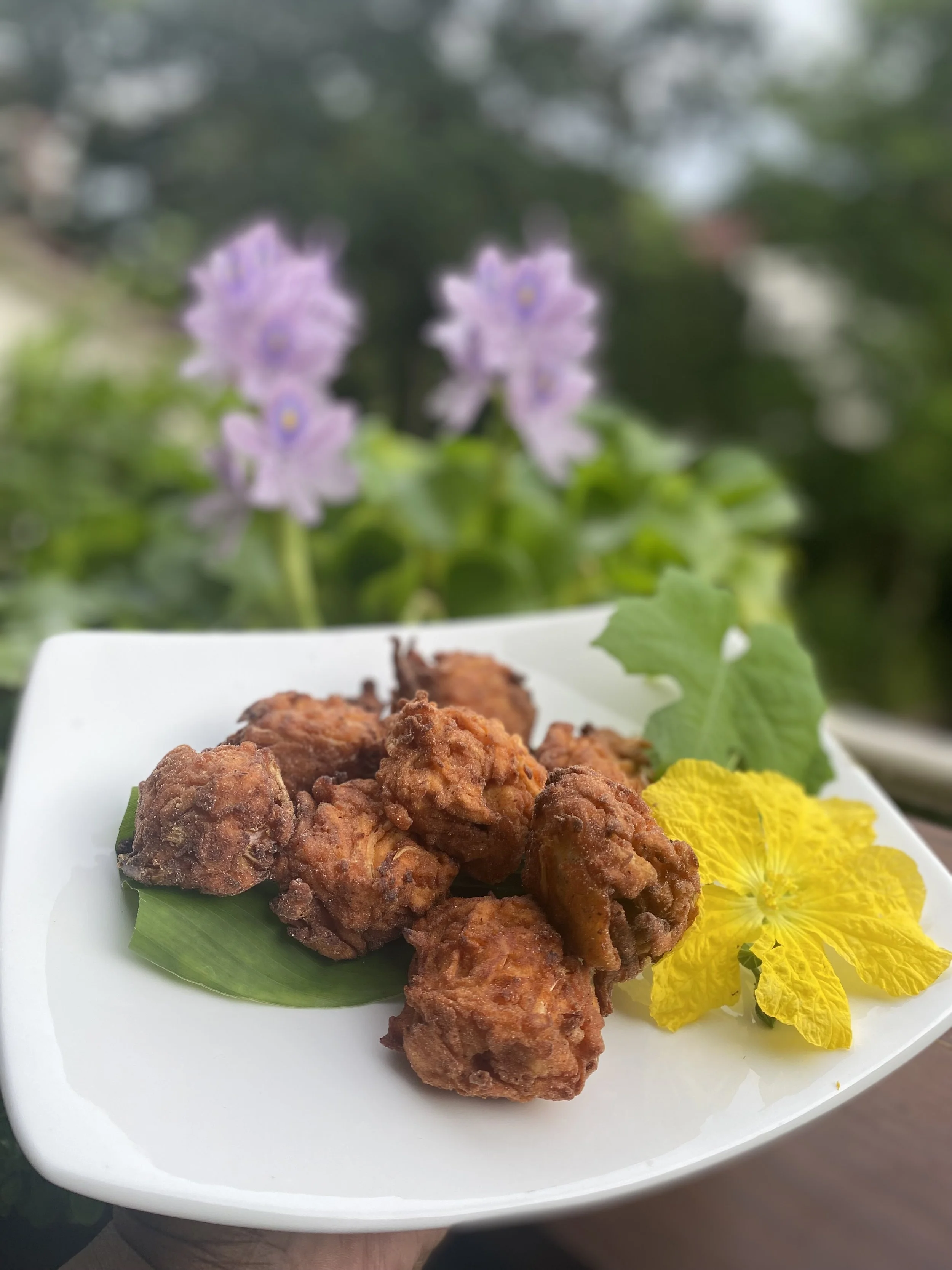 Fried chicken pieces on a white plate with green leaf and yellow flowers, outdoors with purple flowers and greenery in the background.