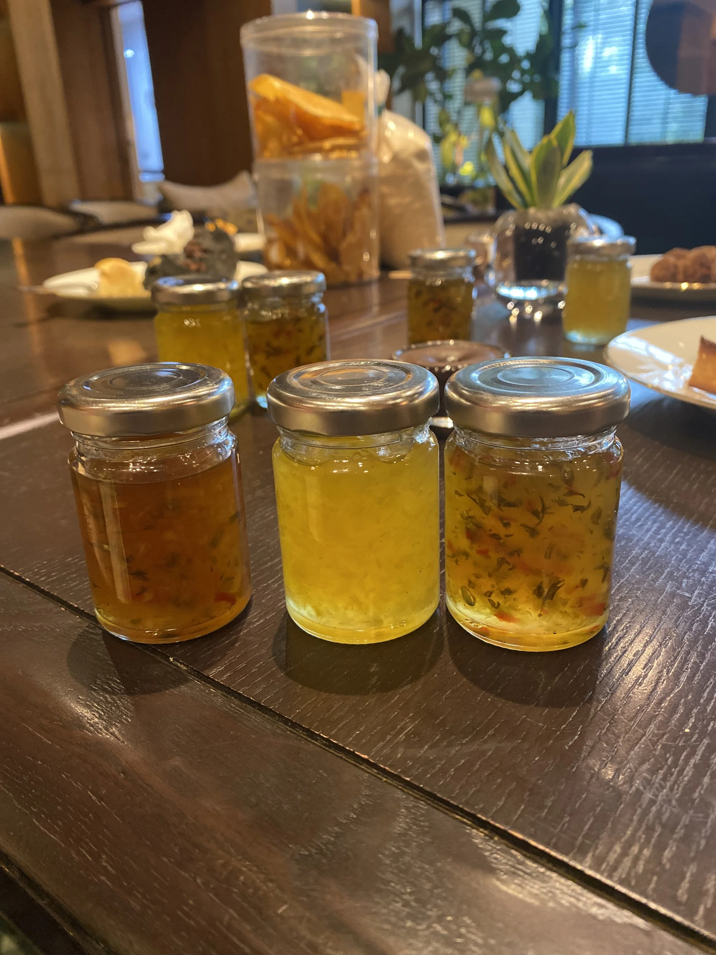 Several small jars of homemade preserves or jams on a wooden table, with other jars and plates in the background.