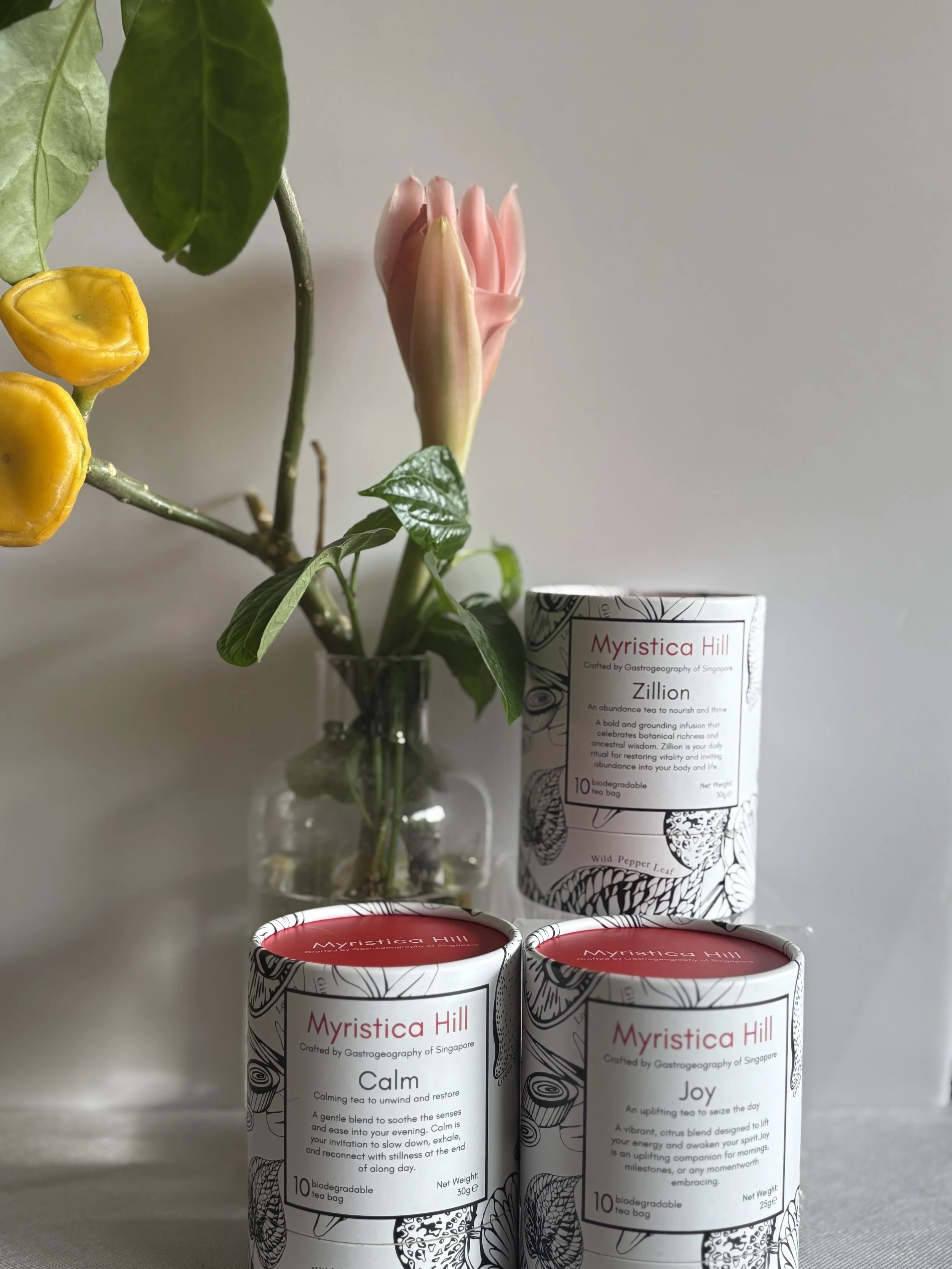 A clear glass vase with green leaves and pink and yellow flowers, placed behind three cylindrical tea packages with black and white floral patterns and red labels, on a white surface against a plain background.