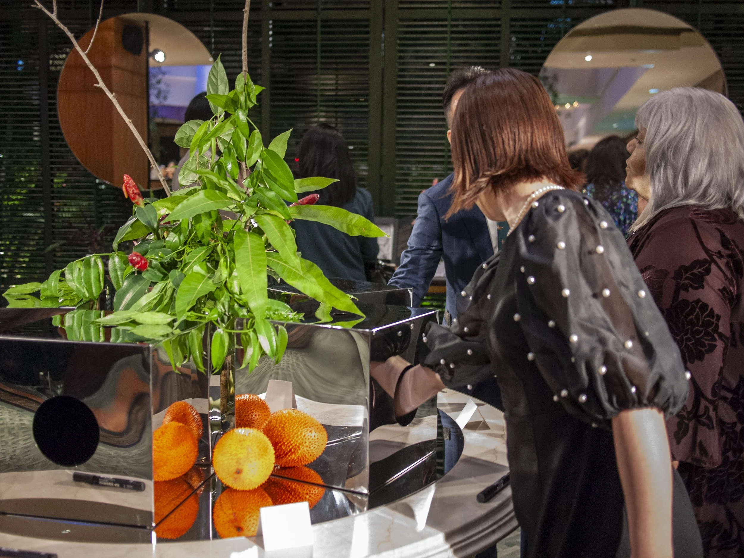People gathered around a piano with a plant and tropical fruit decorations in a stylish indoor setting.