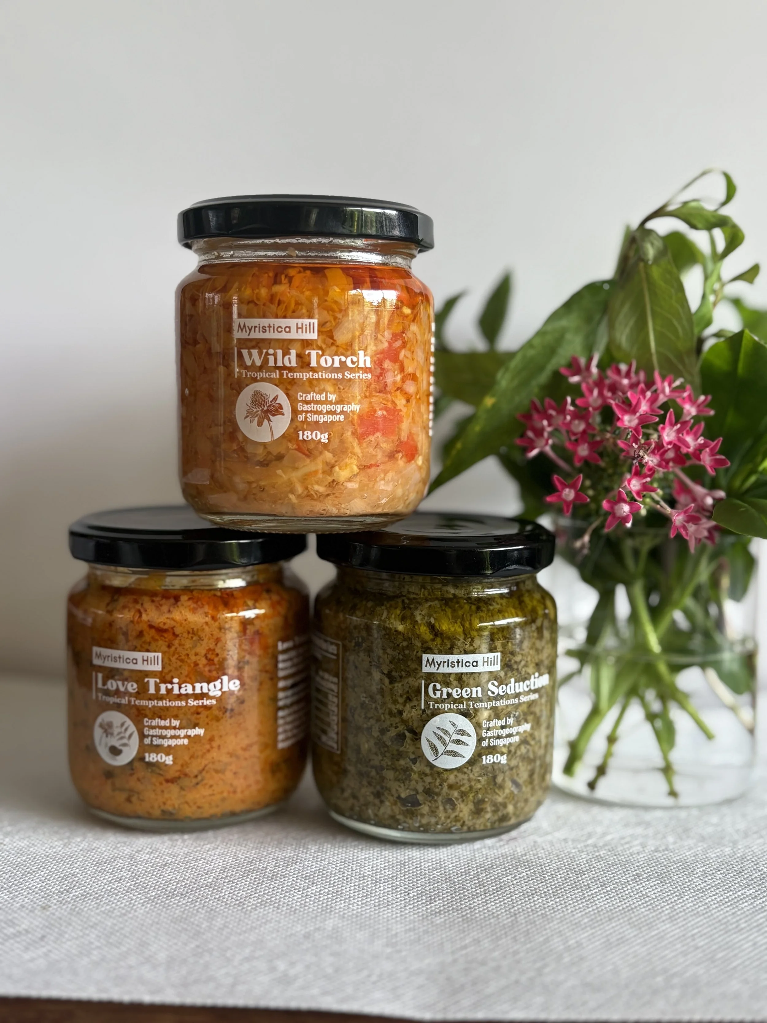 Three jars of artisanal seasonings labeled Wild Torch, Love Triangle, and Green Seduction, with a pink flowering plant in the background, placed on a white tablecloth.