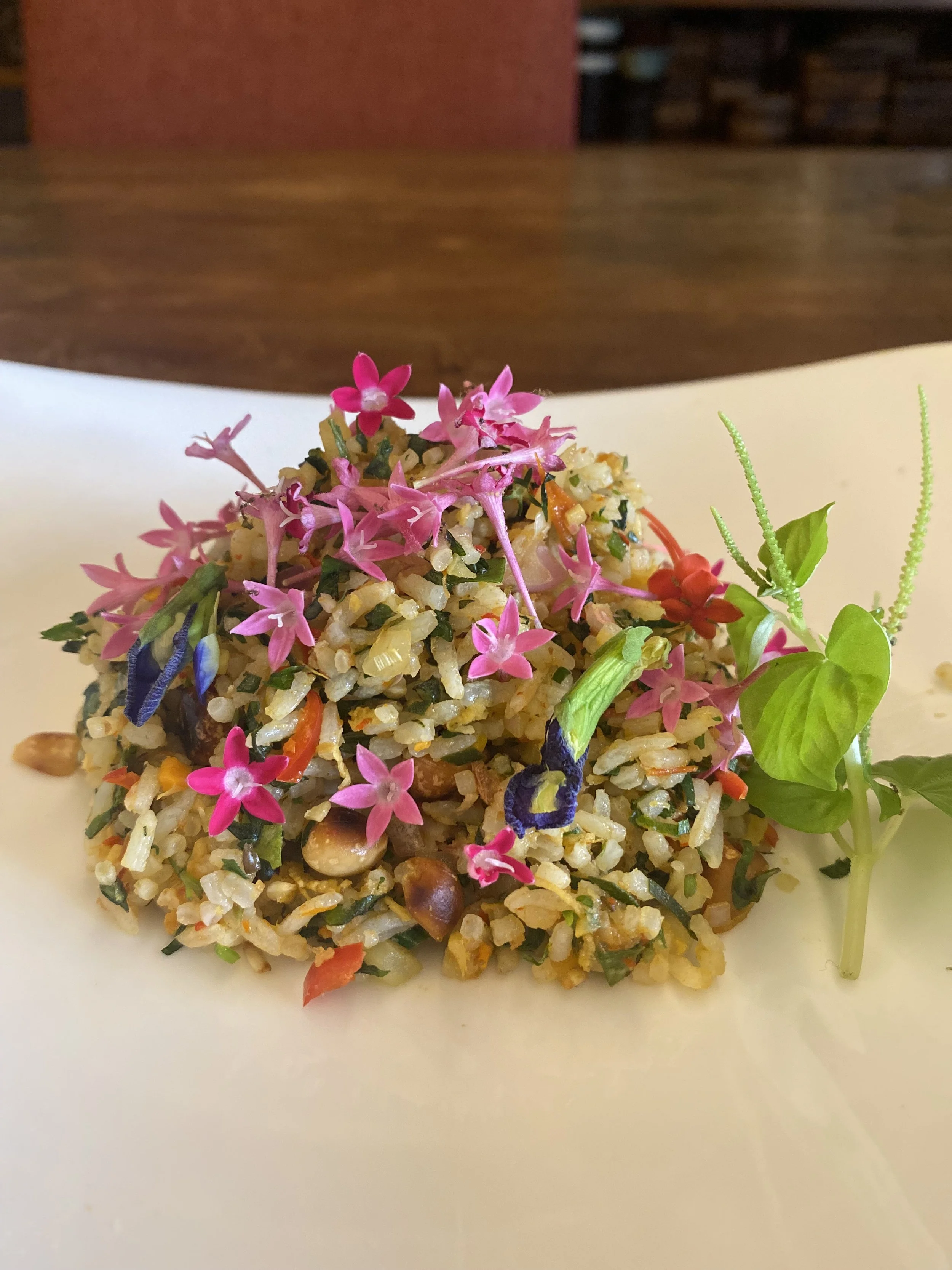 A plate of seasoned rice with chopped vegetables, garnished with pink and purple edible flowers and fresh herbs.