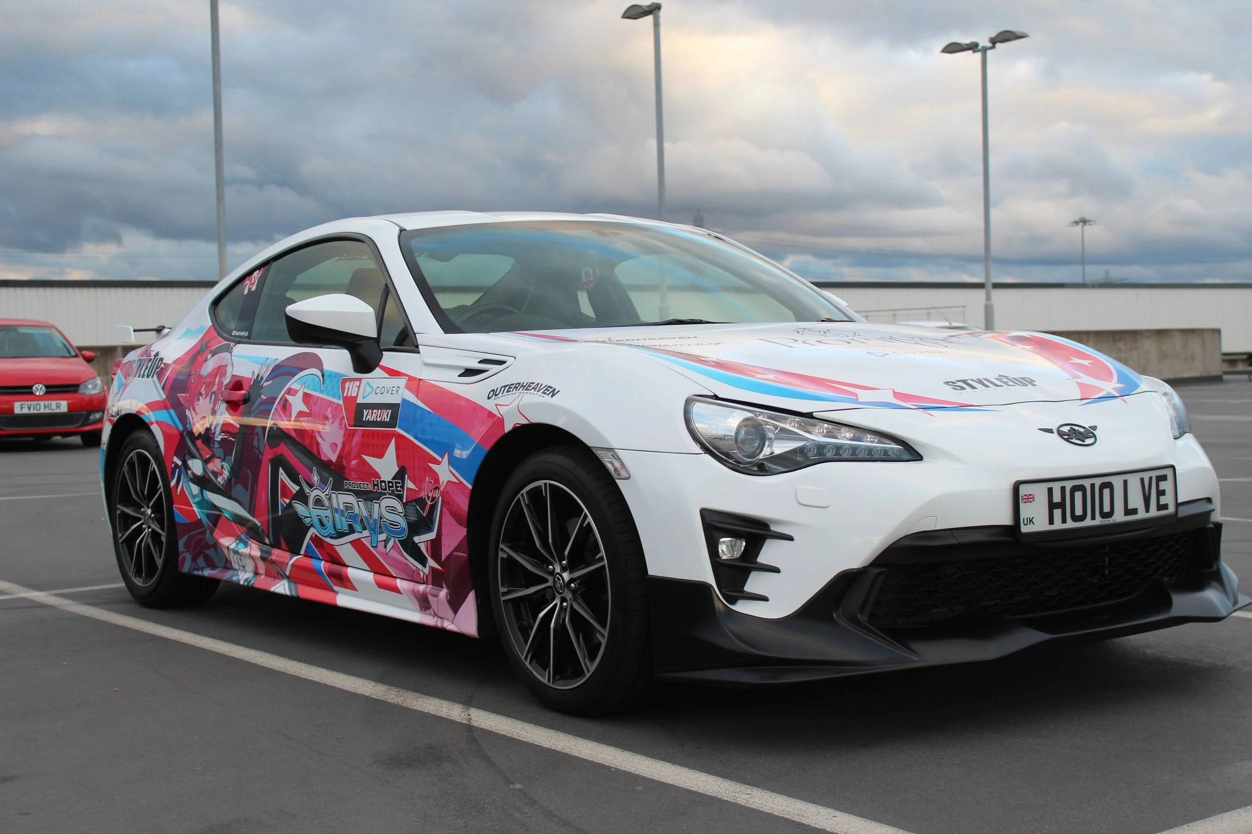 A white sports car with colorful anime-themed decals parked in a parking lot, with other cars and a cloudy sky in the background.