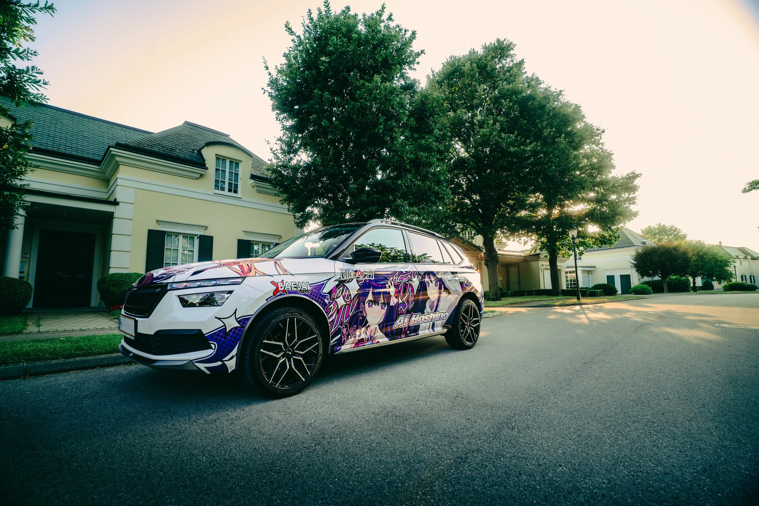 A white SUV with anime-themed decals, including illustrations of a purple-haired girl, parked on a quiet residential street with houses and trees.