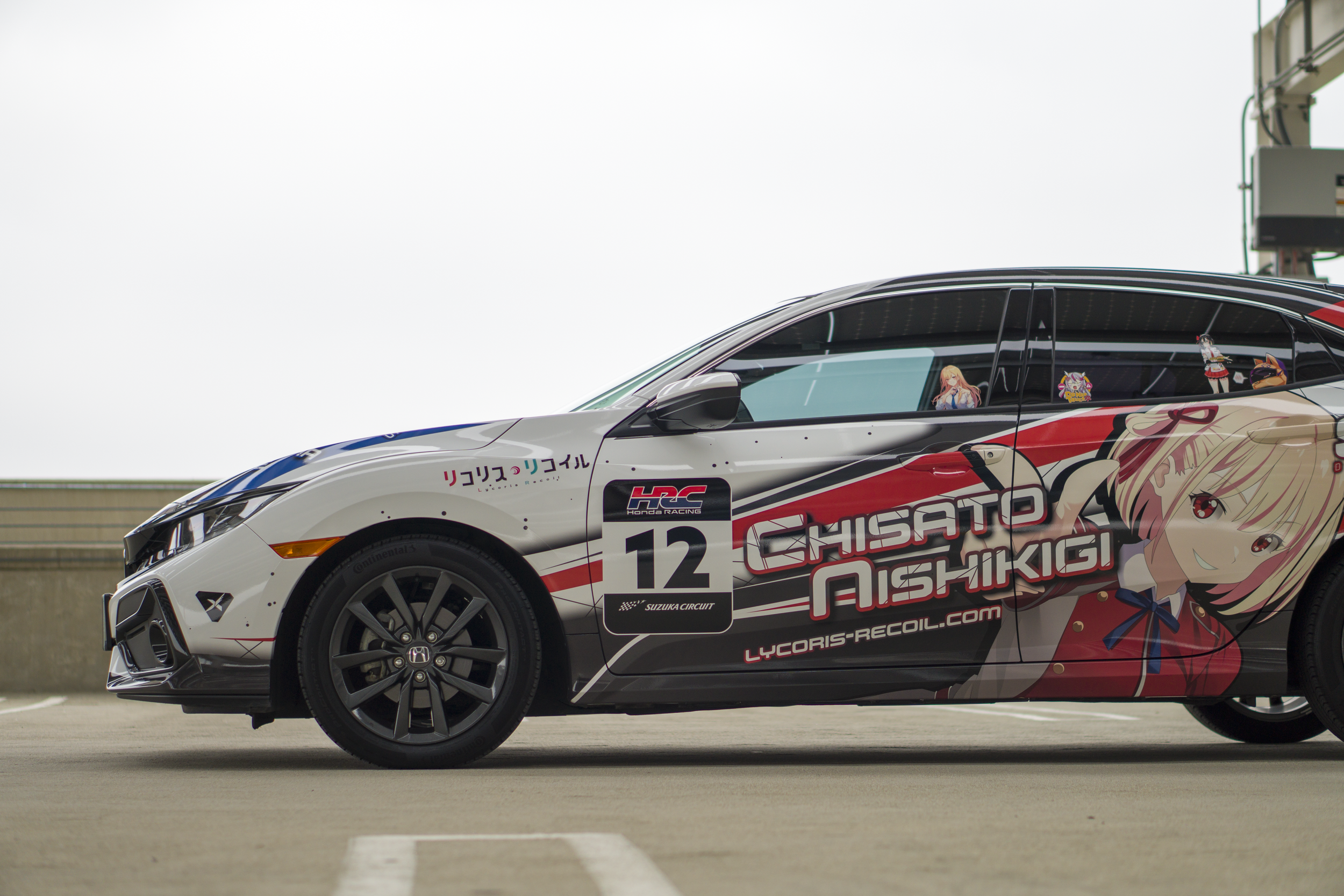 A racing car with anime-themed graphics, including an illustration of a girl with blonde hair on the side and various stickers, parked on a racing circuit.