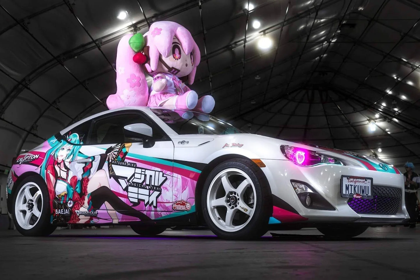 A white sports car with colorful anime graphics, including Hatsune Miku, on the side, is parked under a large, arched metal structure. A large plush doll of My Little Pony character Pinkie Pie is sitting on the car's roof. The car's headlights have p