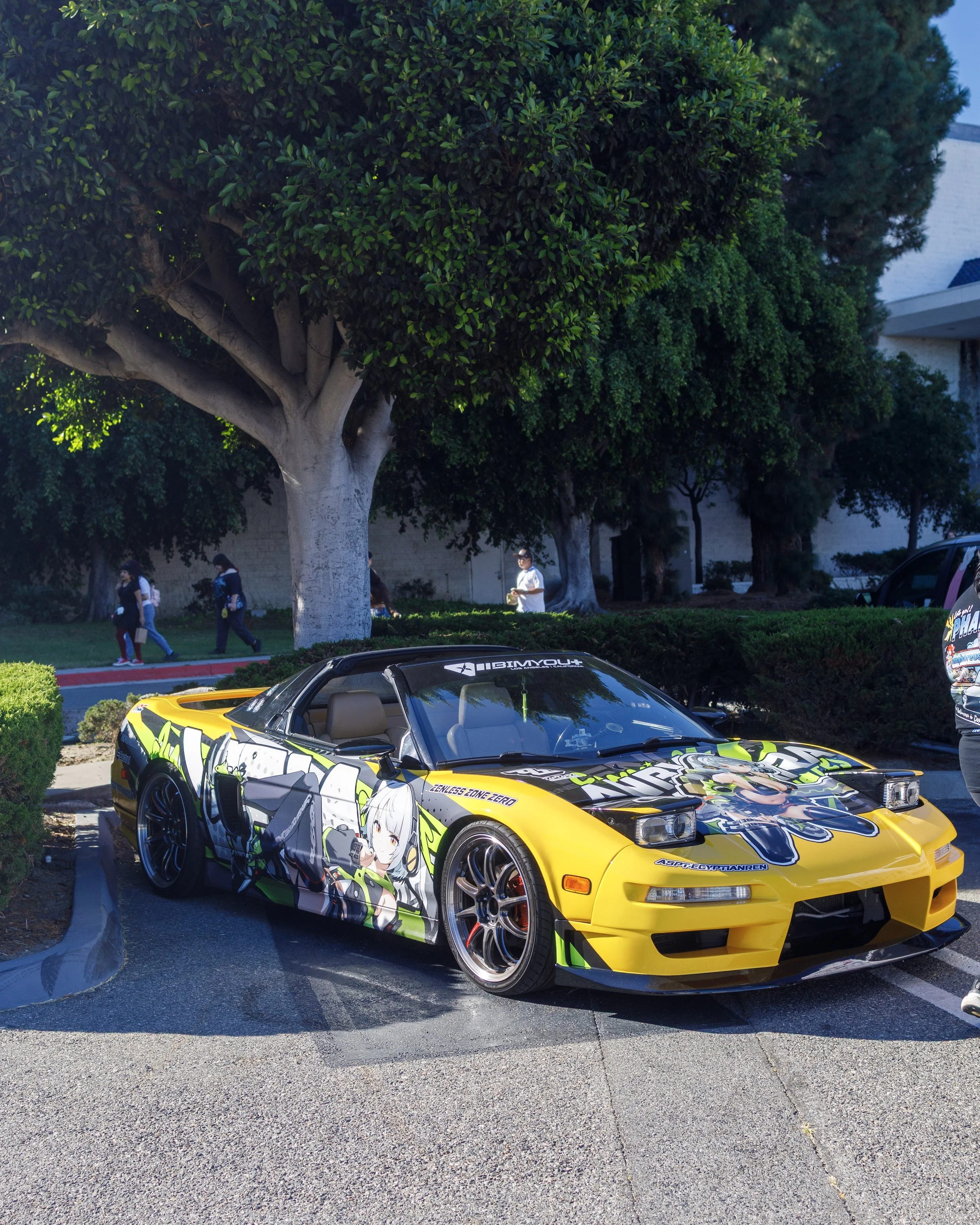 A yellow sports car with anime character graphics parked under a large tree on a parking lot, with people walking in the background.