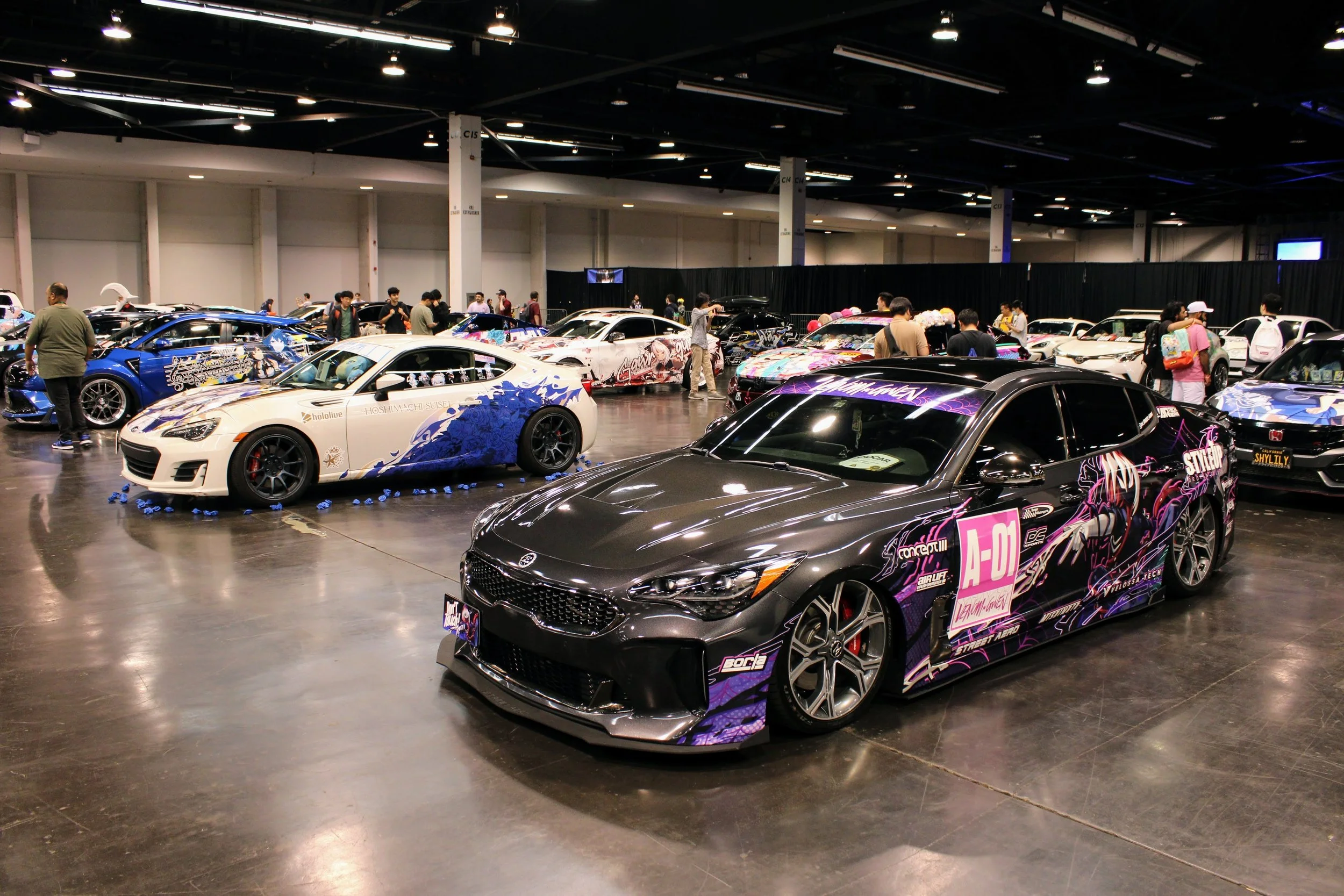 Showroom filled with modified sports cars, including a black and purple car with artwork and a white and blue car with blue flame designs, surrounded by attendees at a car show.