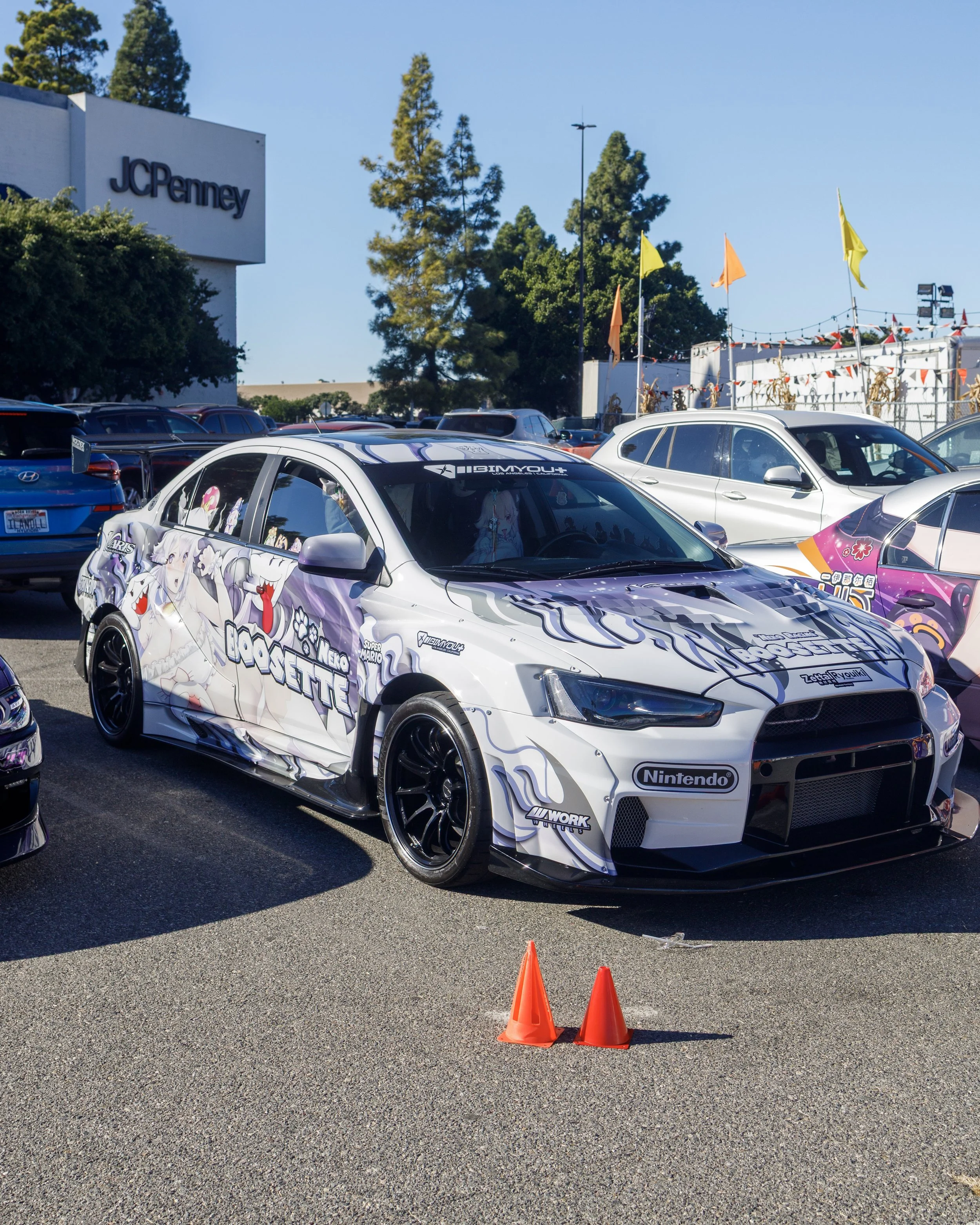 A white racing car with anime-style artwork and sponsorship logos, parked in a lot with other cars, in front of a JCPenney store, with small orange traffic cones in front.