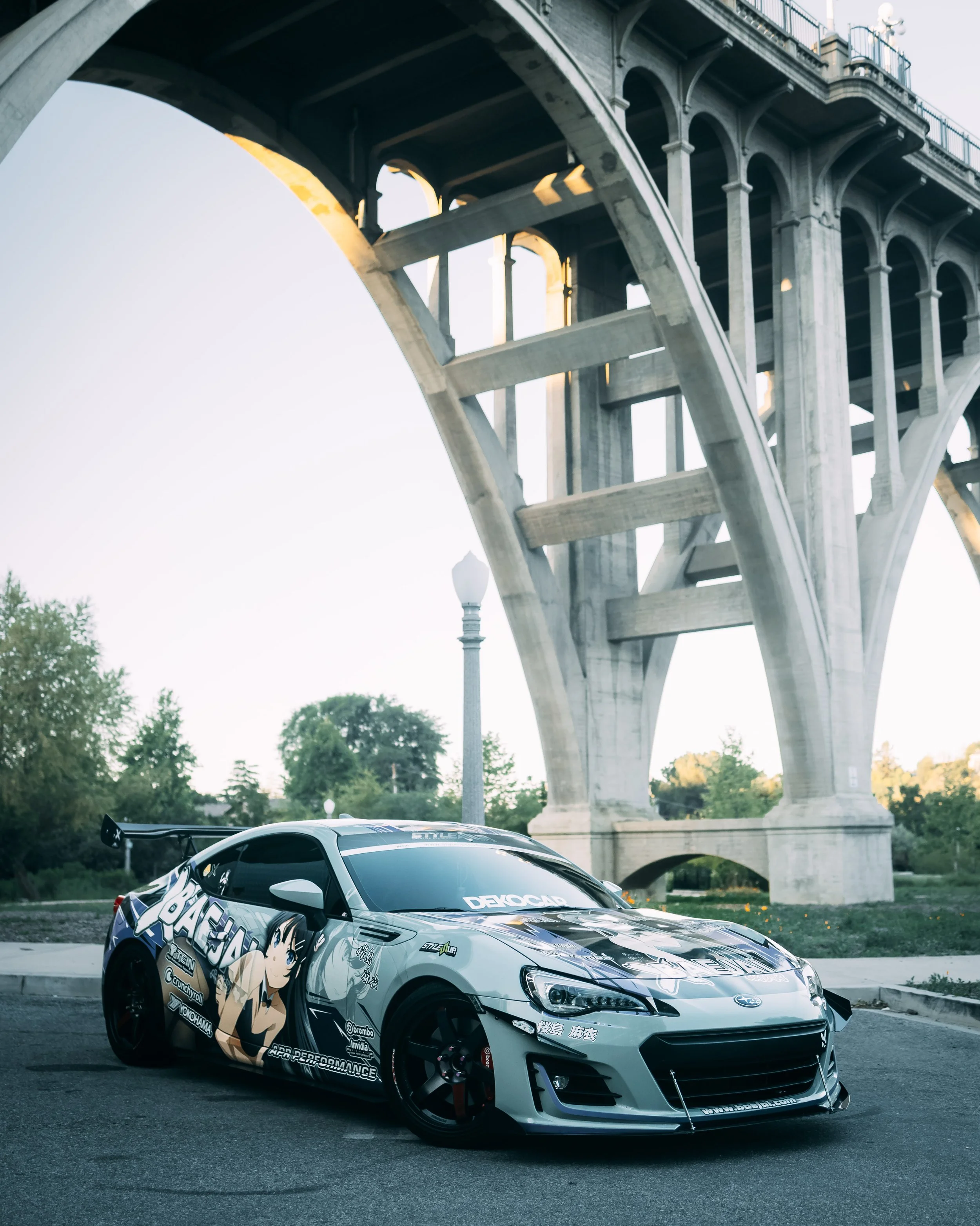 A silver sports car with anime-style graphics parked under a large bridge during daylight.