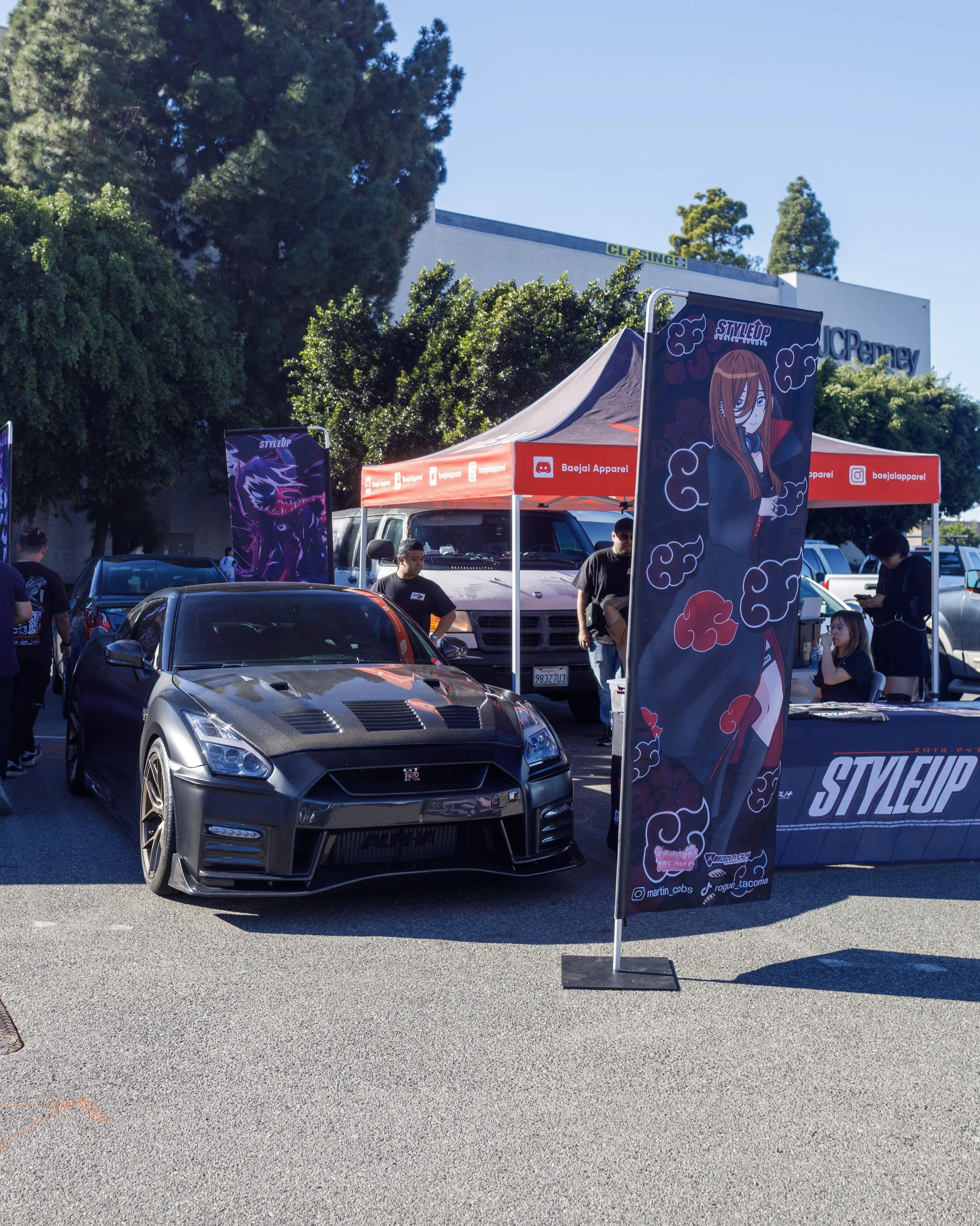 A black Nissan GT-R parked in front of a booth at an outdoor car event, with banners displaying anime-style artwork and a red canopy tent labeled 'Baejali Apparel' and 'STYLEUP'.