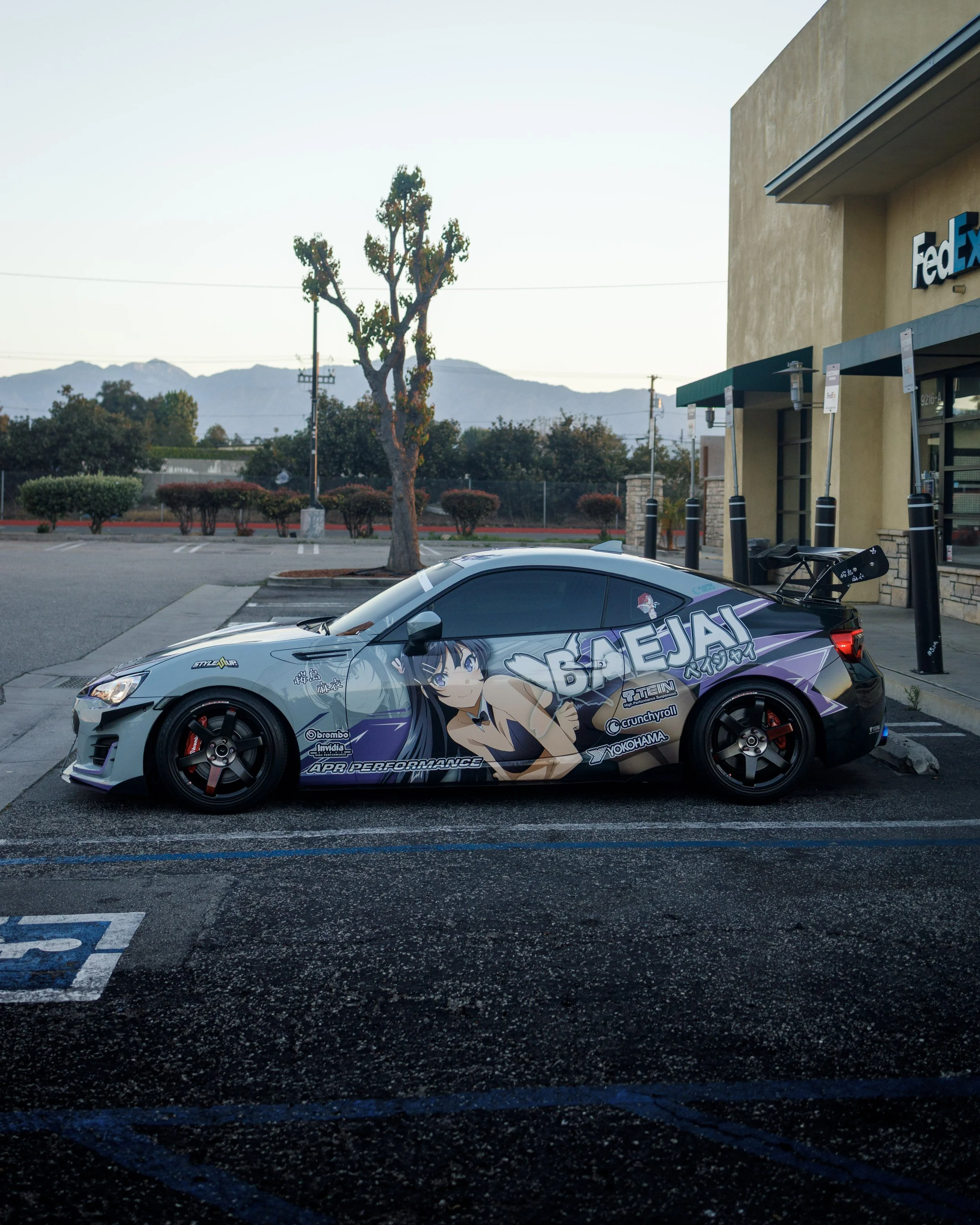 A sports car with anime-style graphics and sponsorship logos parked outside a FedEx store in a parking lot, with mountains and trees in the background.