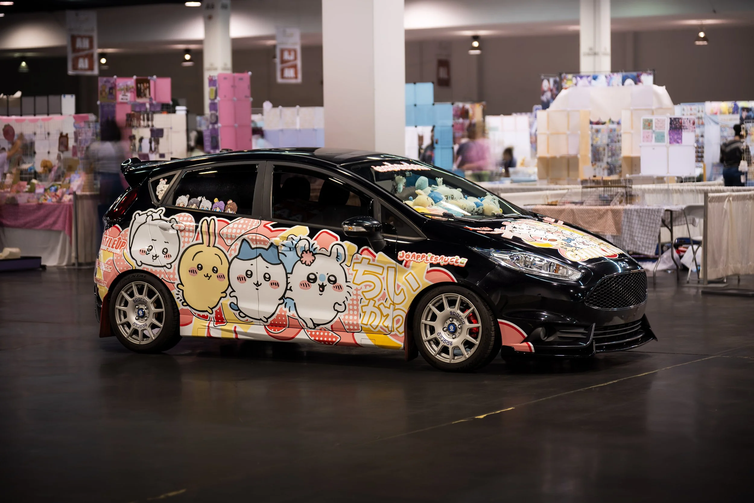 Black car decorated with colorful anime-style characters and Japanese text, displayed indoors at an event with booths and tables in the background.