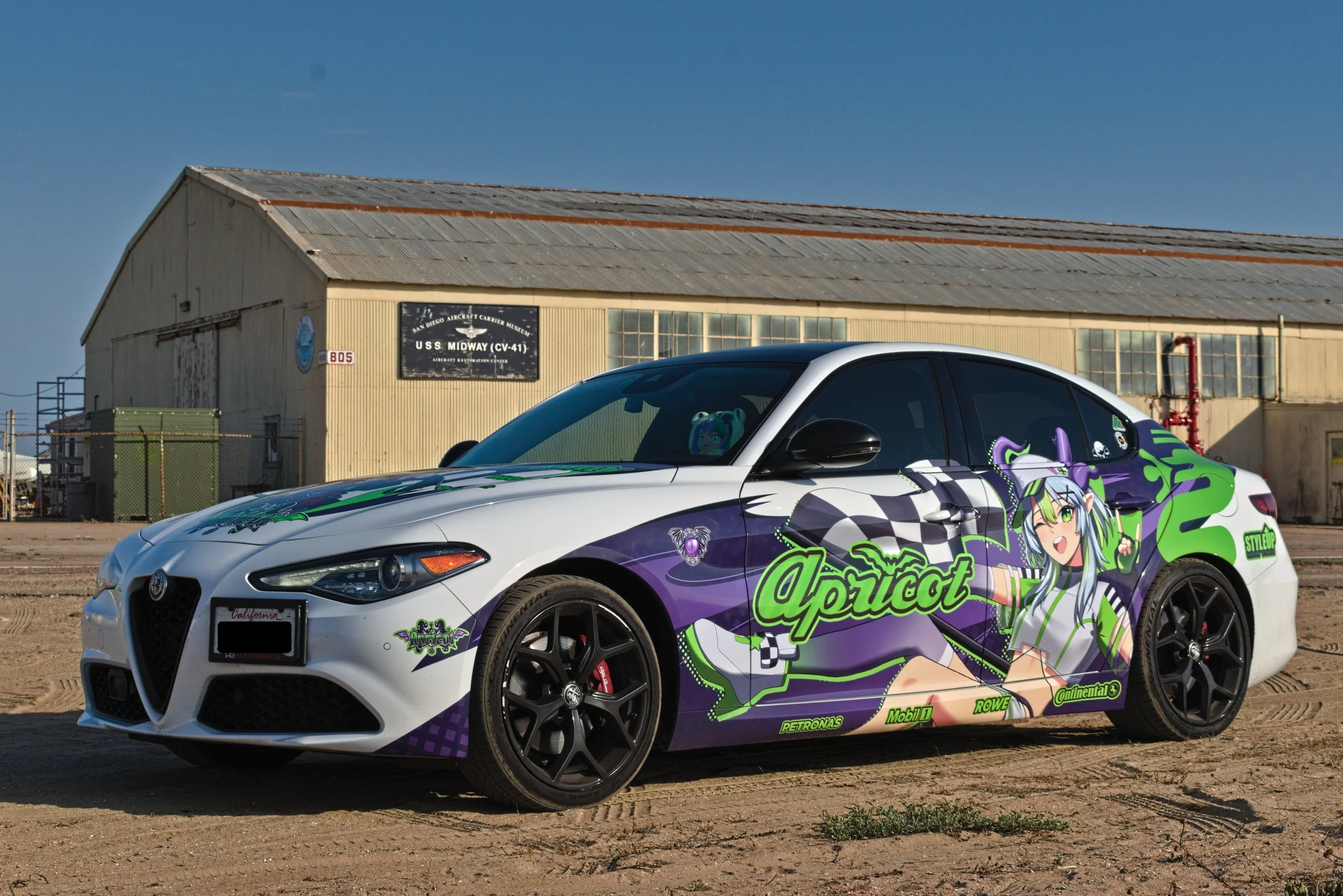 A white sports car with colorful anime-style decals and the word 'apricot' on the side, parked on a dirt lot near an old building.