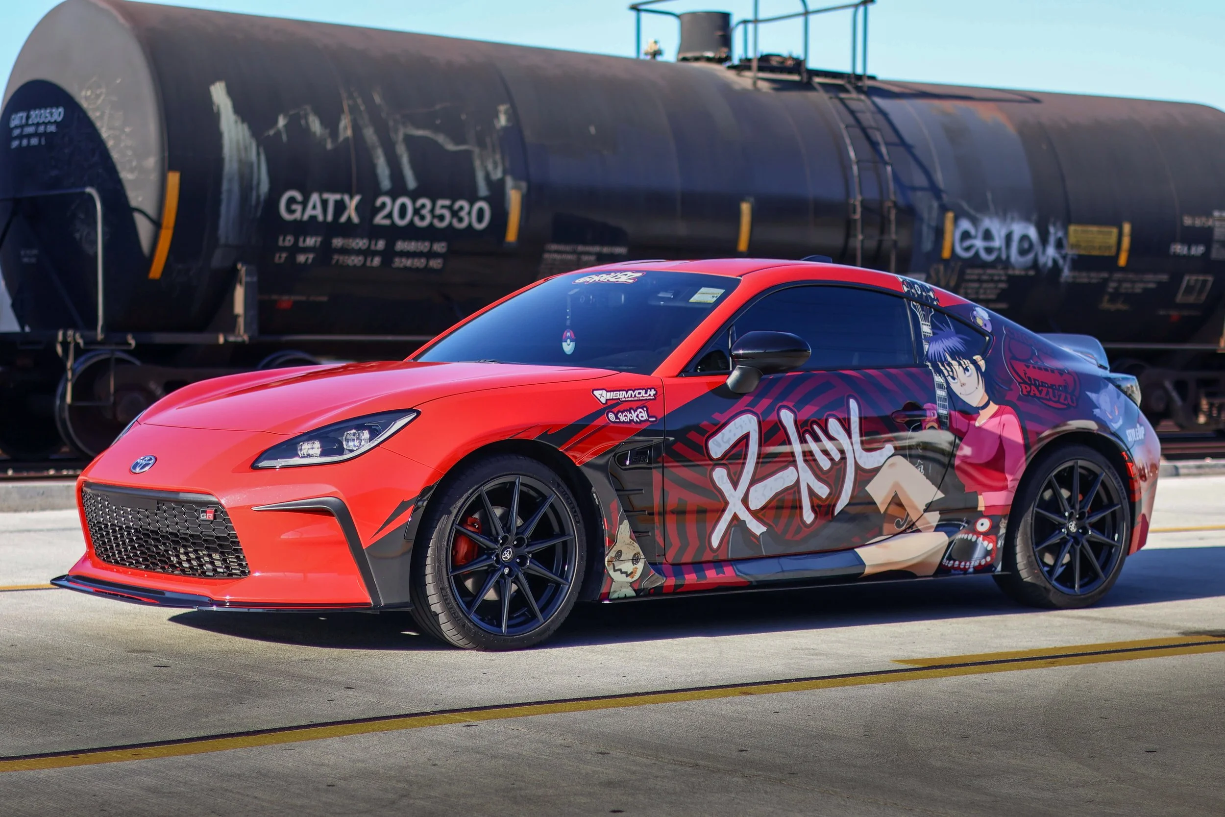 A red sports car with custom anime-style graphics on its side parked on a road, with a large black tanker train in the background.
