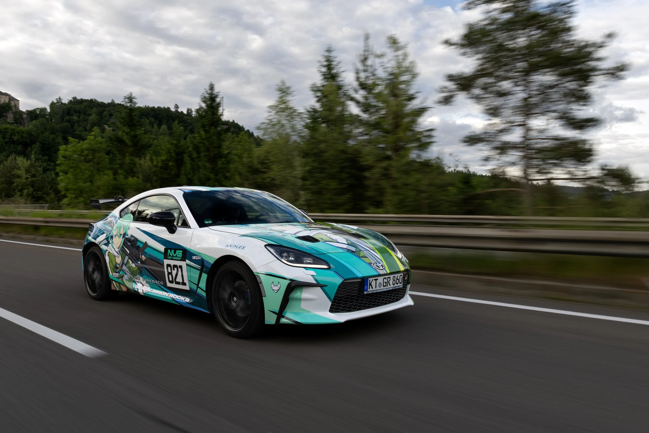 A white sports car with anime-themed decals racing on a highway with a wooded background under a cloudy sky.