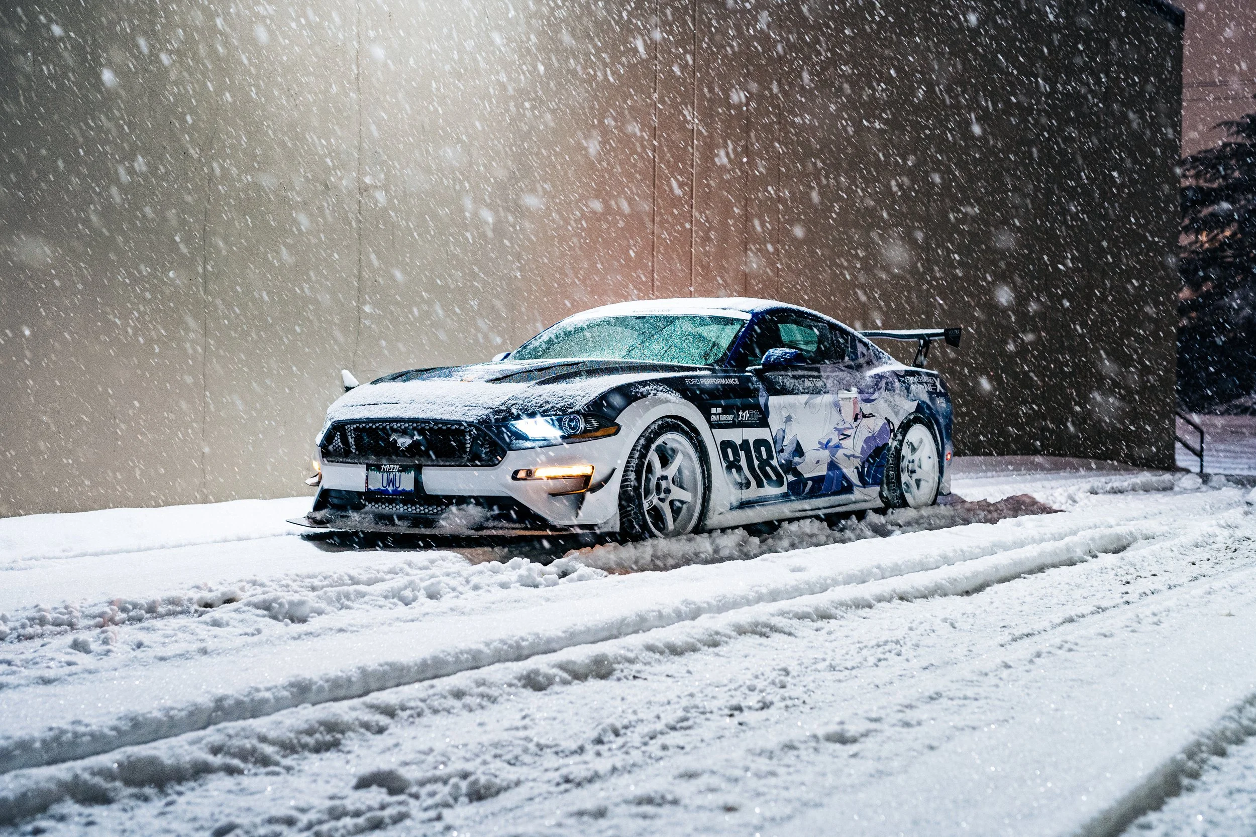 A race car with racing decals and the number 818 is parked in the snow during a snowstorm at night.