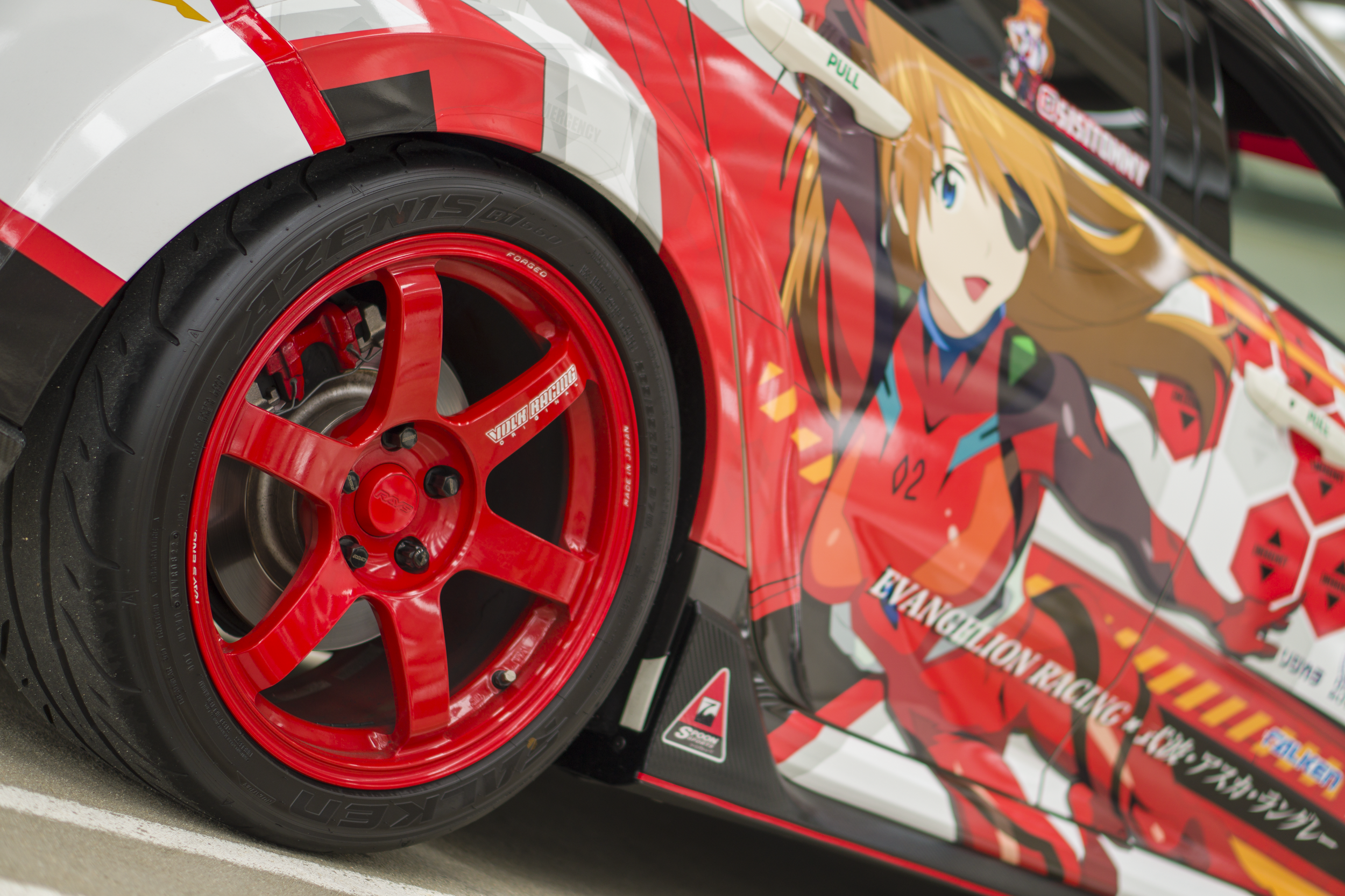 Close-up of a race car's front wheel and side panel featuring an anime character from Evangelion with brown hair, blue eyes, and a red and black racing suit.