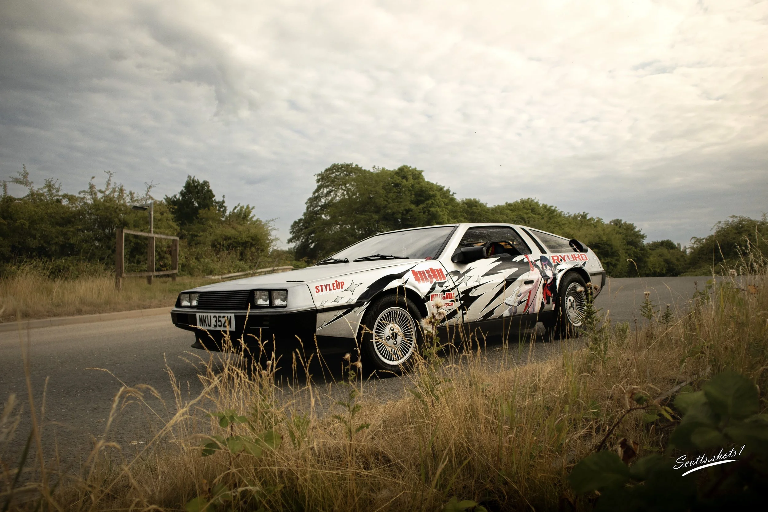 A white sports car with anime-style graphics and red text parked on the side of a rural road with grass and trees in the background under a cloudy sky.