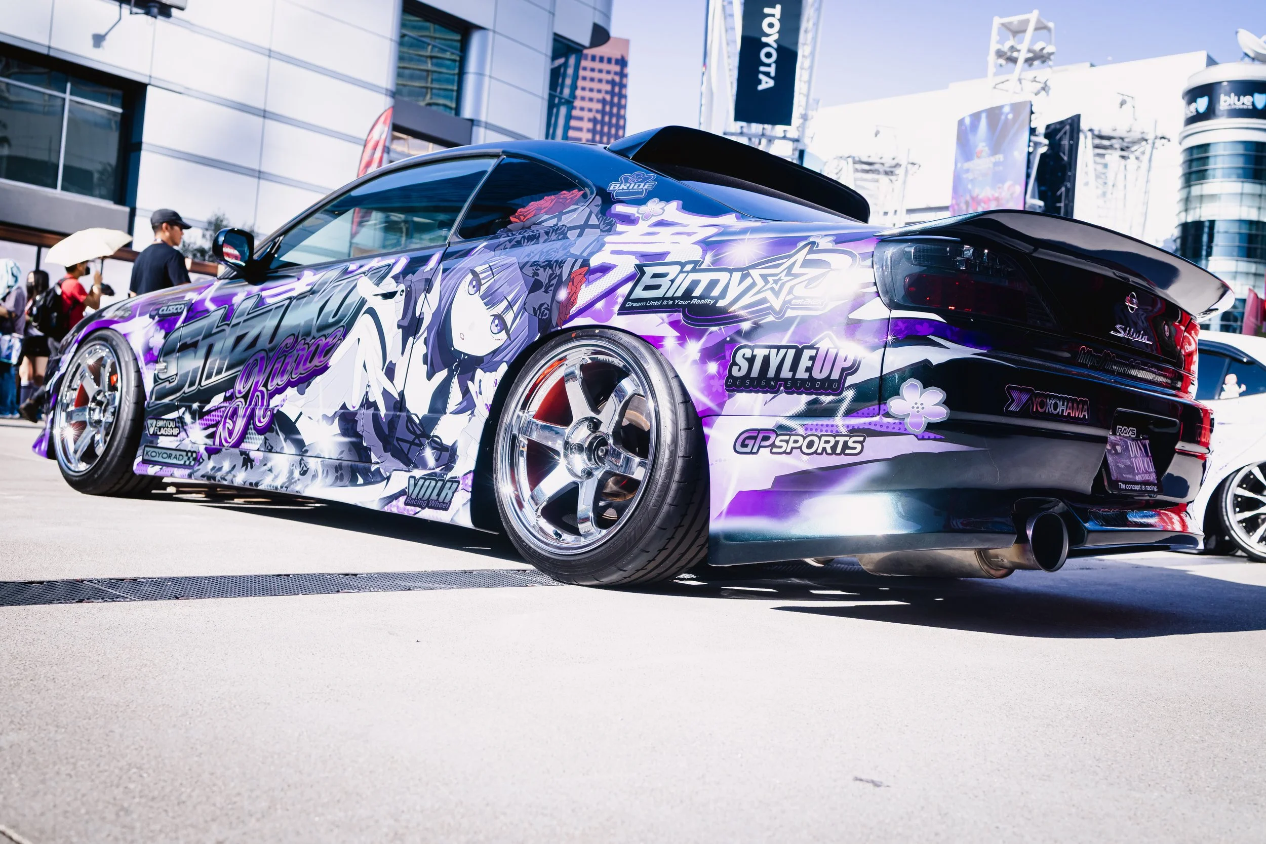 A modified sports car with a vibrant anime-themed wrap featuring a female character, at an outdoor car event. The car has custom wheels, low-profile tires, and various sponsor decals, with a crowd and buildings in the background.