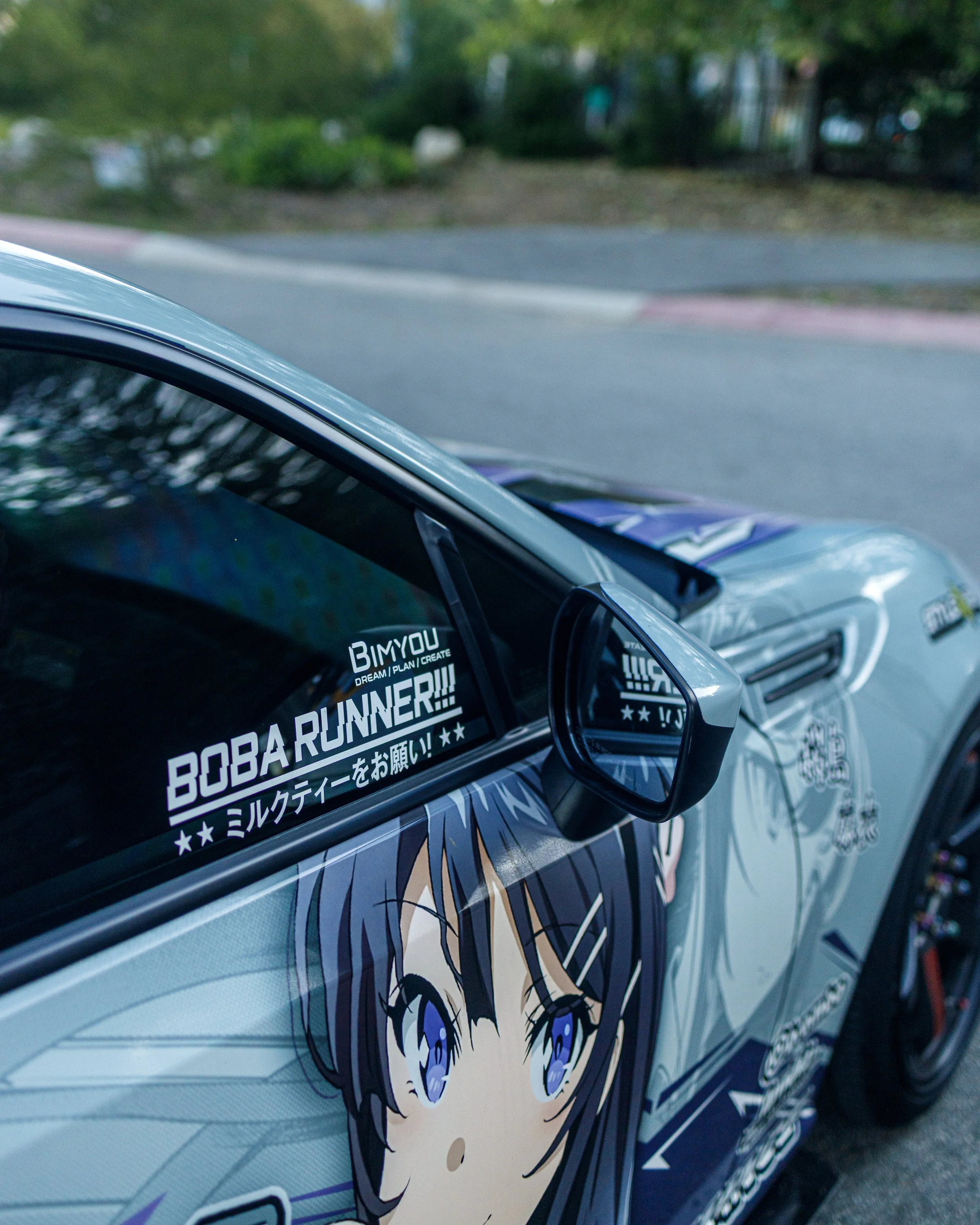 A race car with anime-style graphics and a side mirror, parked on a street with green trees and sidewalk in the background.