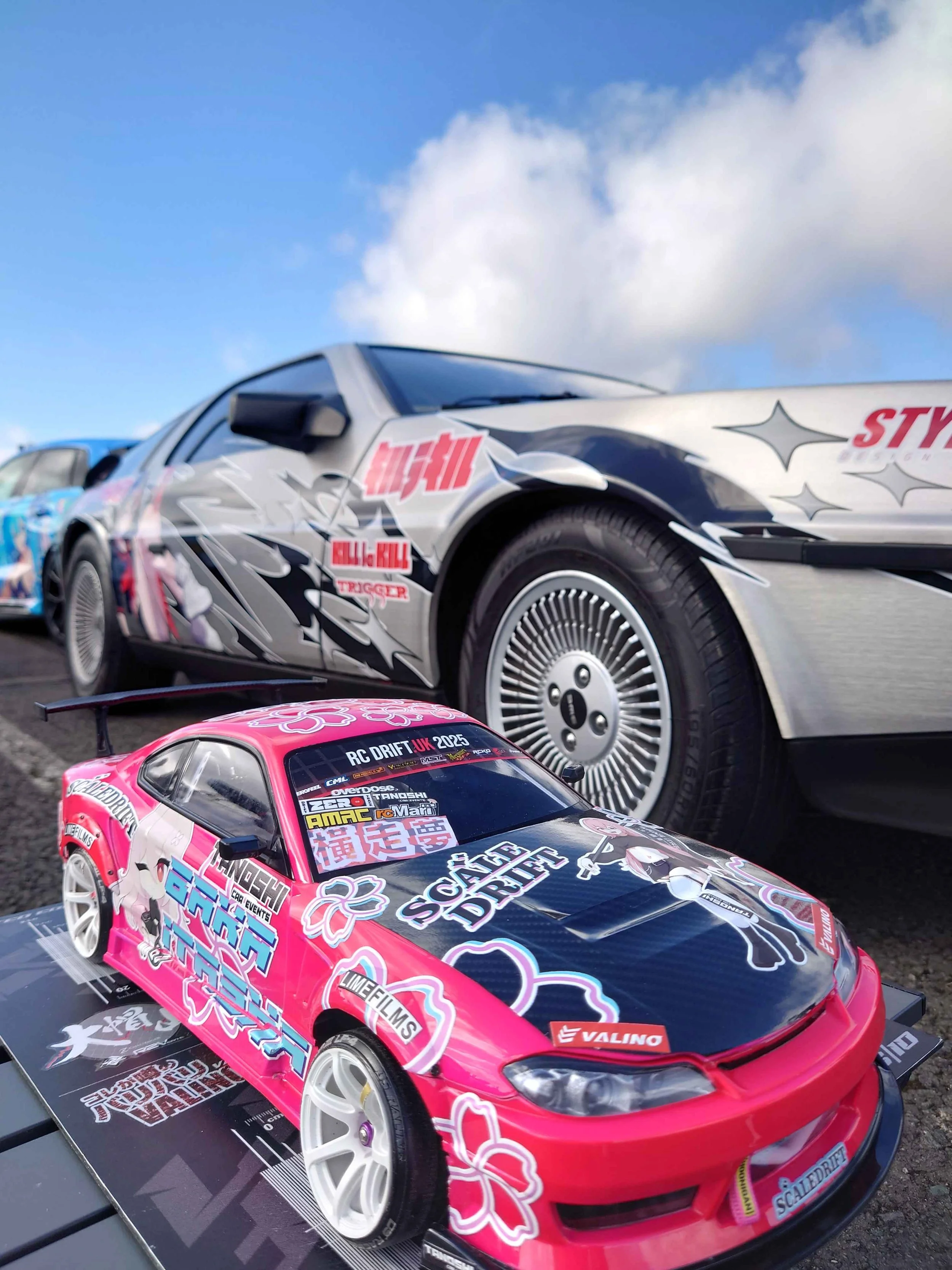 A pink and black remote control drift car modeled after a sports car with decals and stickers, placed outdoors on a stand, with a classic silver sports car with custom graphics in the background and a partly cloudy blue sky overhead.