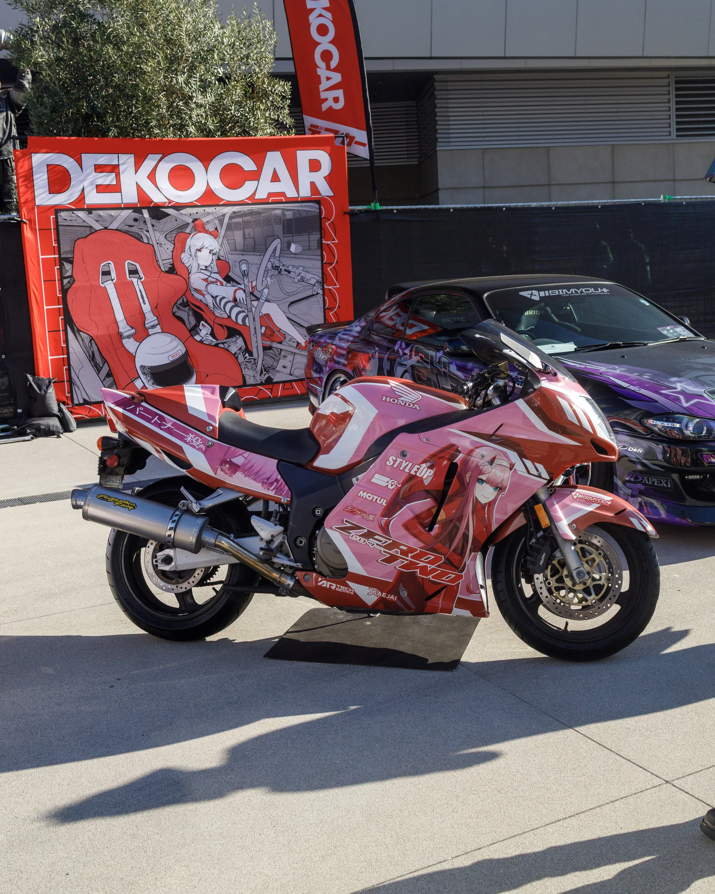 A pink motorcycle with anime-style character artwork on the fairings, parked outdoors with cars and a large red Dekocar banner and artwork display in the background.