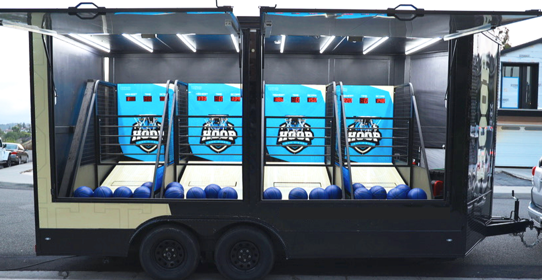 Mobile mini bowling alley with three lanes and blue balls in black trailer with logo, set up outdoors.