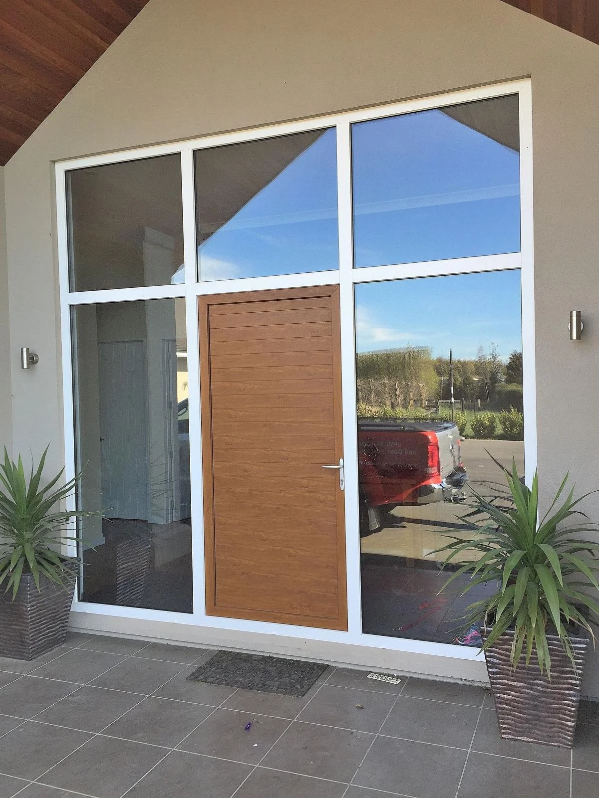 Modern house entrance with large glass windows, a wooden door, and potted plants on either side