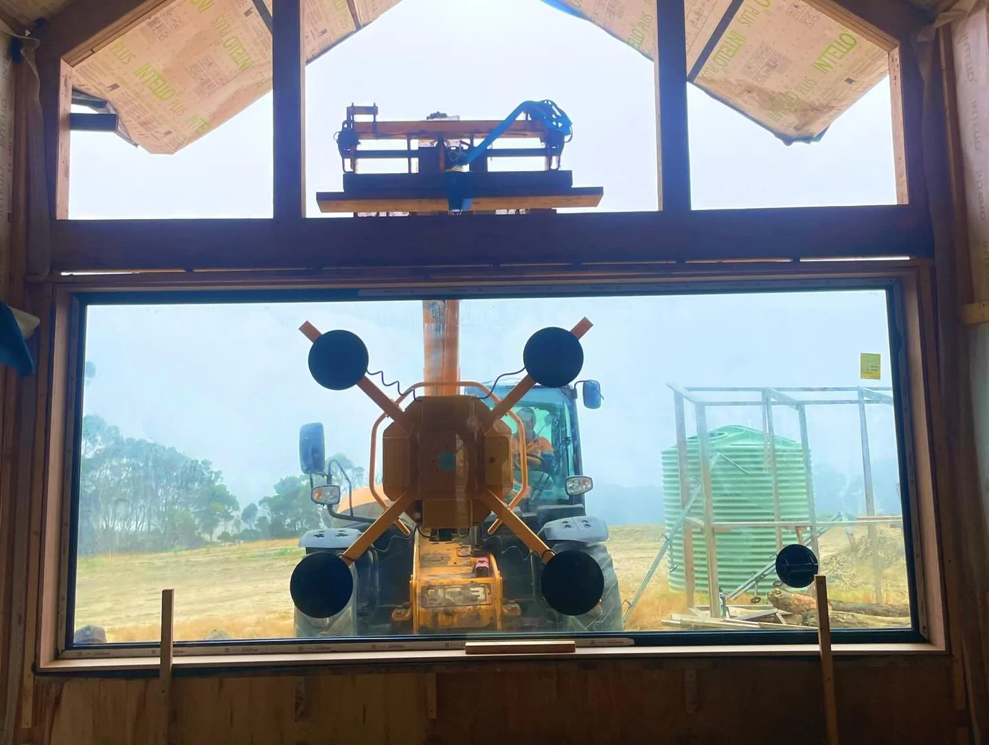 A spectacular image of a large scale window being glazed on a construction site with a backhoe loader in the foreground and a yellow construction lift above, with trees and overcast sky in the background.