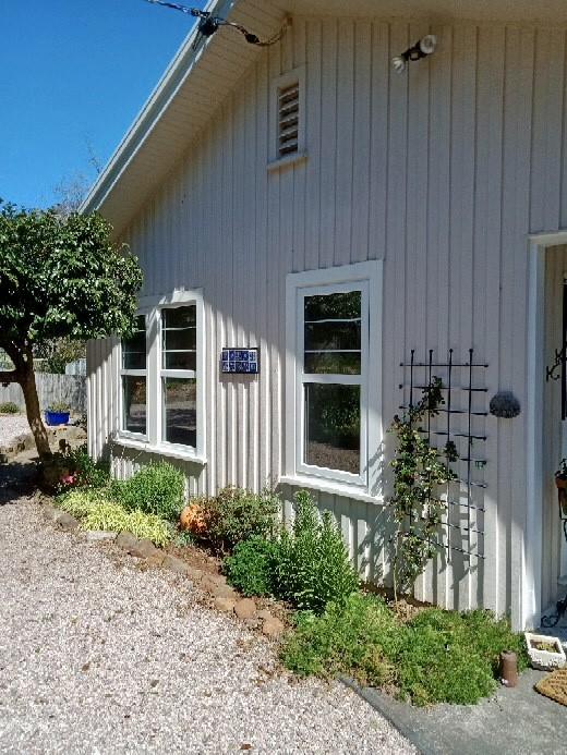 A white house with vertical siding, two white framed windows, a small garden with green plants and a rose bush, a gravel pathway, and a decorative trellis on the exterior wall, under a clear blue sky.