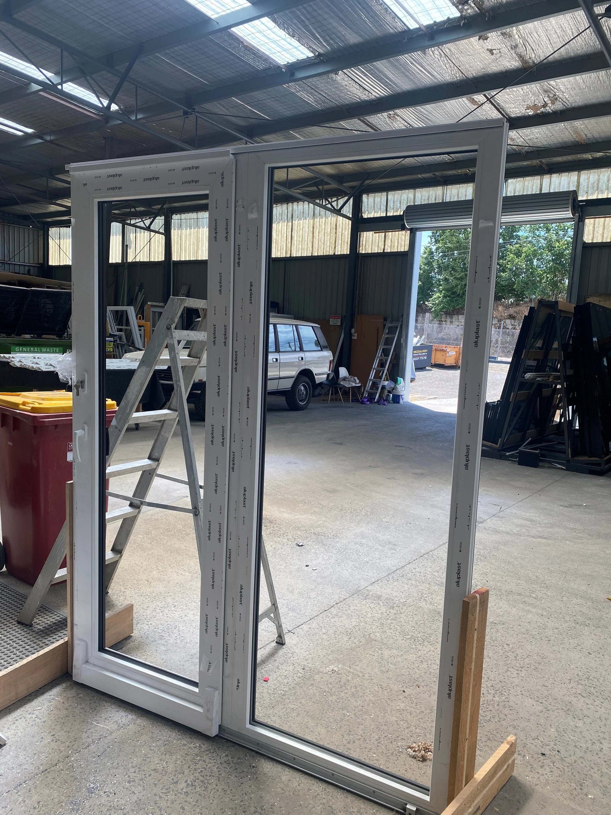 A metal and glass door frame in a warehouse or workshop with tools and a ladder nearby, and a vehicle parked outside visible through the glass.