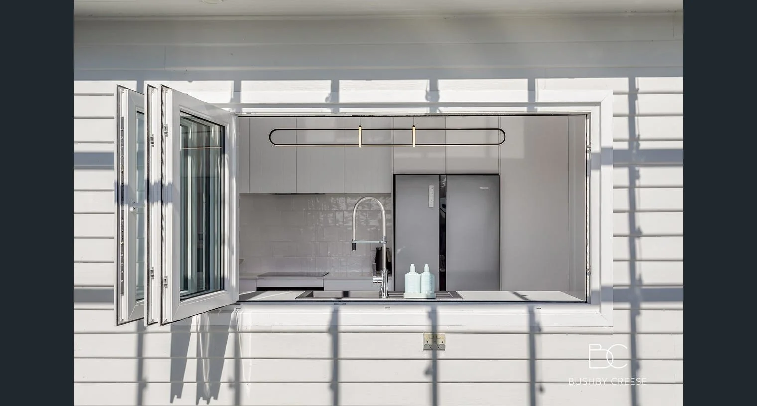 A modern kitchen view through an open window in a white exterior wall. Inside, white cabinets, a gray refrigerator, a black induction stove, and a silver faucet over a sink with three bottles visible. Shadows from the window frame cast across the exterior wall.