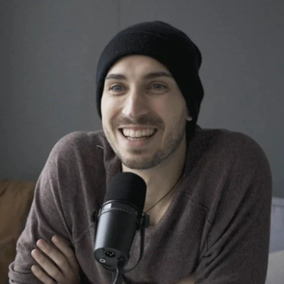 A smiling man wearing a black beanie and a brown shirt, sitting in front of a microphone.