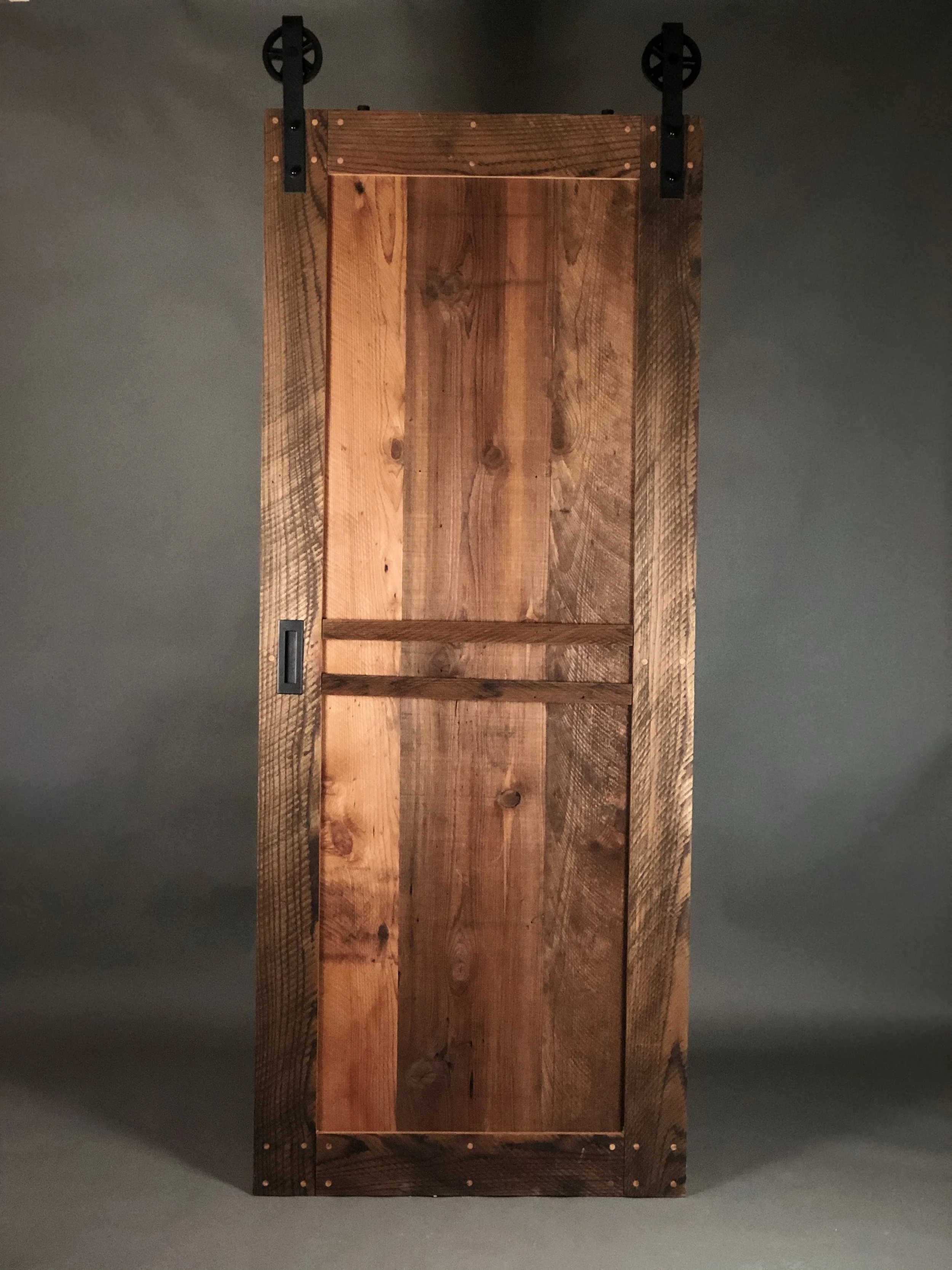 A rustic wooden sliding barn door with black hardware, including wheels at the top, against a plain dark wall background.