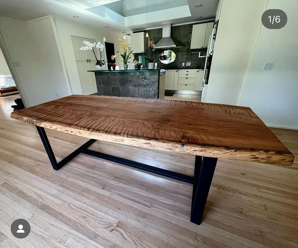 A modern wooden table with a live edge, supported by black metal legs, located in a bright, open kitchen area.