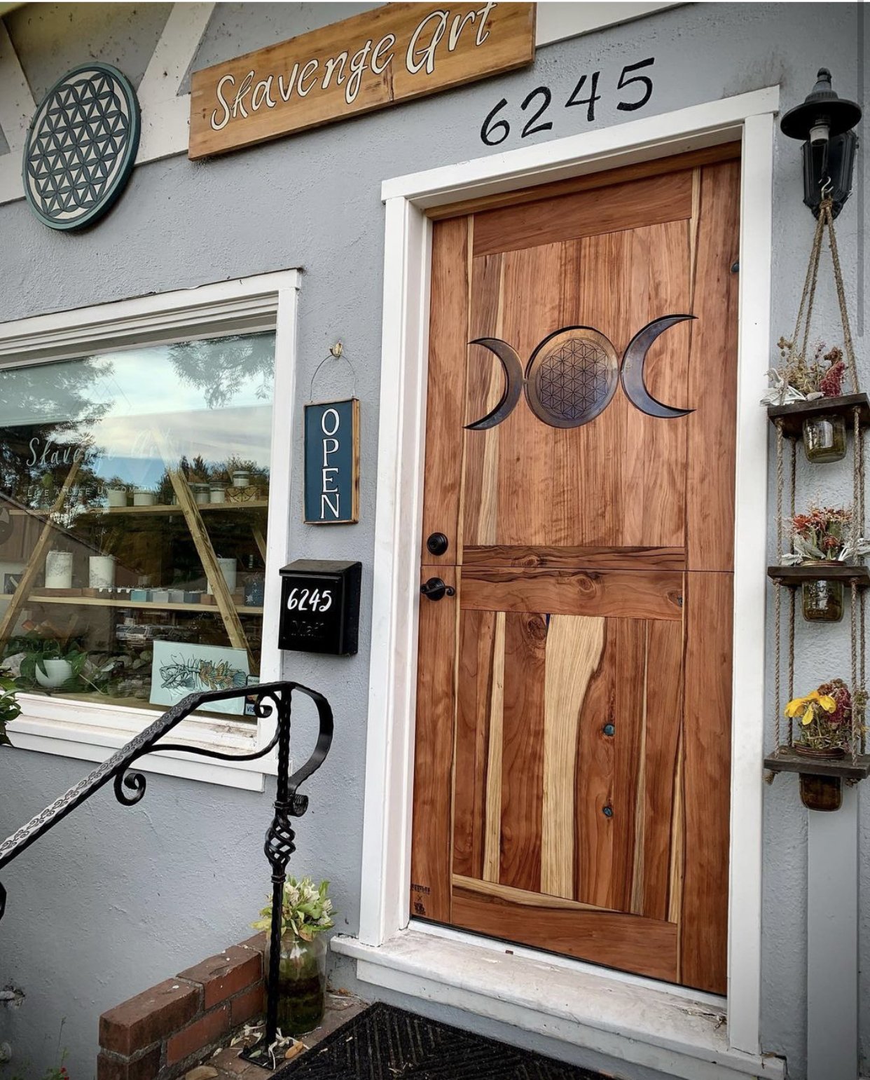 Wooden front door with decorative moon phases and sacred geometry symbol, blue 'OPEN' sign, house number 6245, window with plants and art inside, exterior wall decorated with small plant shelves, black mailbox, iron railing, and potted plant at the e