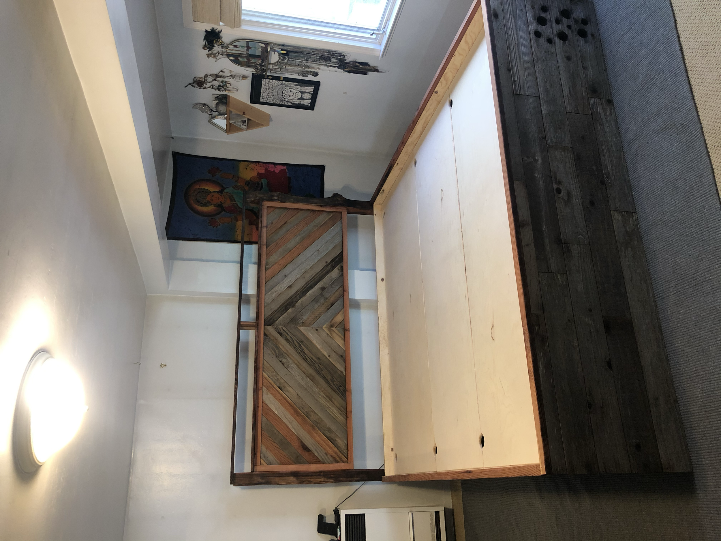 Empty wooden table with a two-tone wooden headboard behind it, decorated with colorful artwork and religious images on a white wall, near a window with blinds.