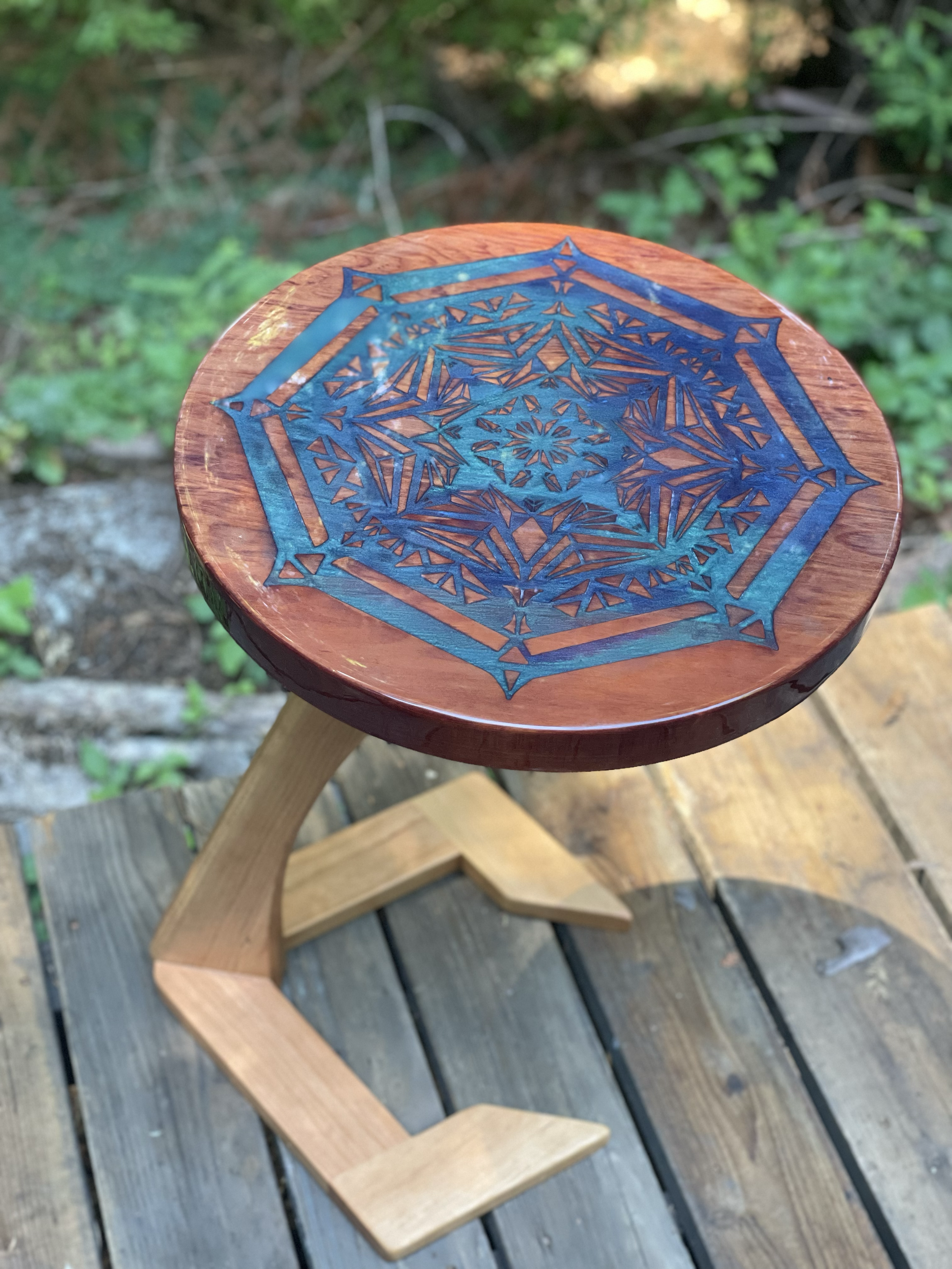 A small wooden stool with an intricate blue geometric design on the round seat, placed outdoors on a wooden deck with greenery in the background.