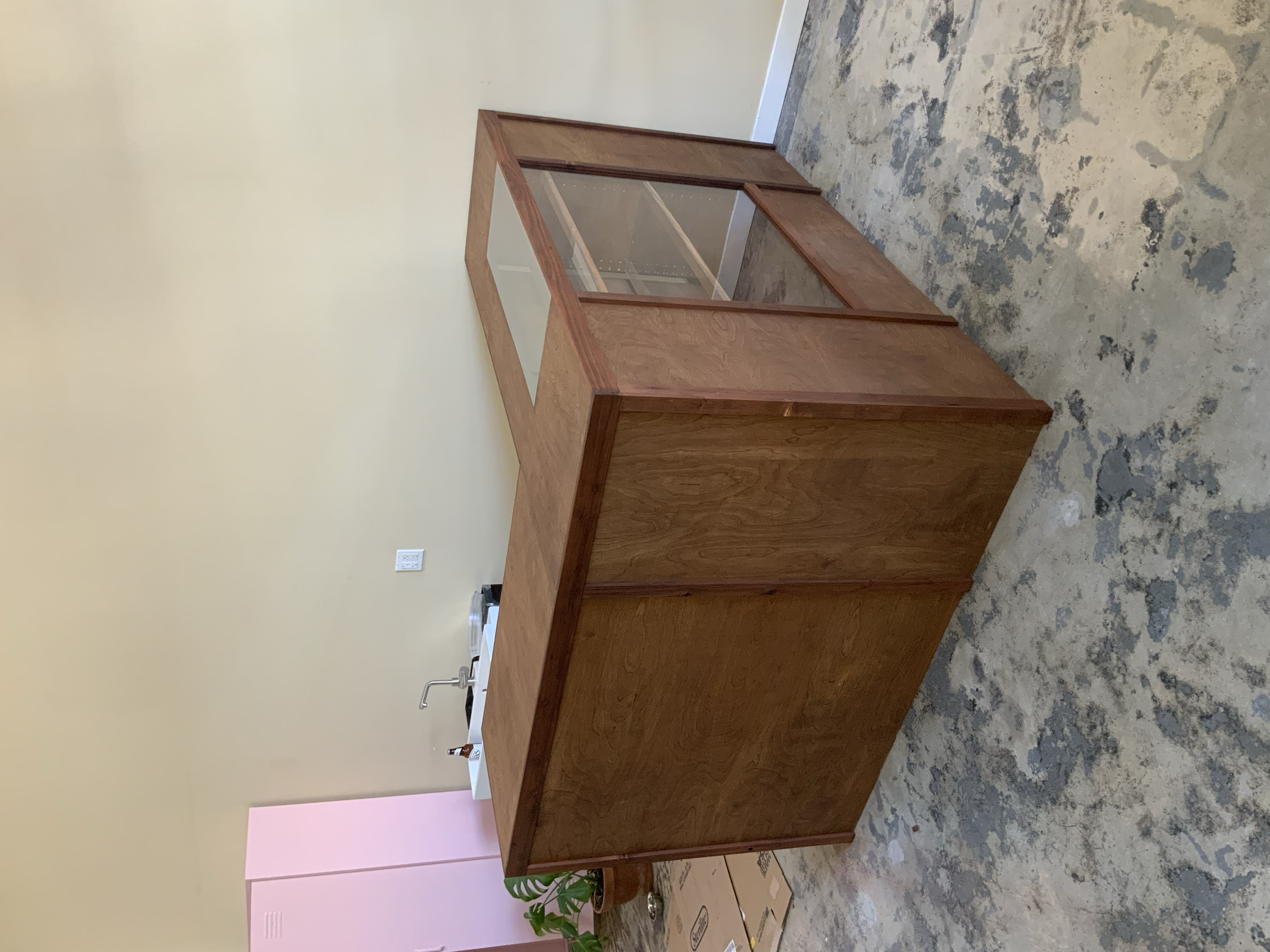 Unfinished wooden cabinet with glass doors, sitting on a concrete floor in a room with yellow walls and a pink section in the corner.