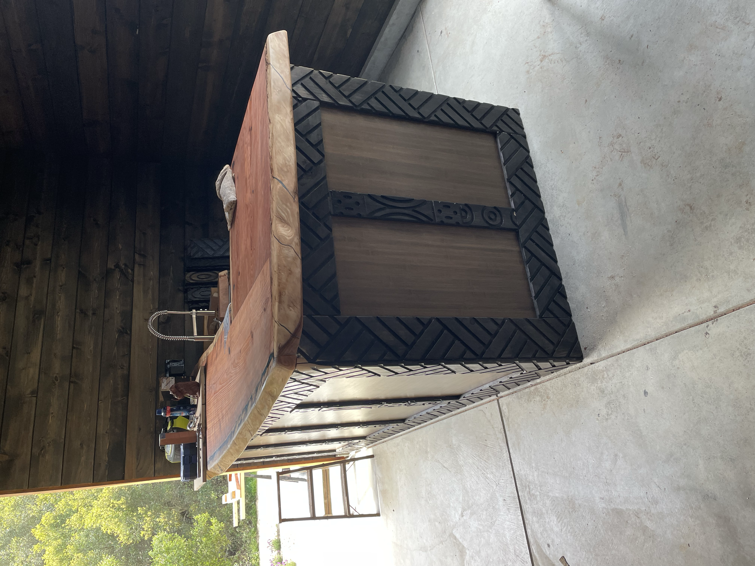 Outdoor kitchen island with wooden countertop, black patterned trim, and a sliding door, located on a concrete patio with trees in the background.
