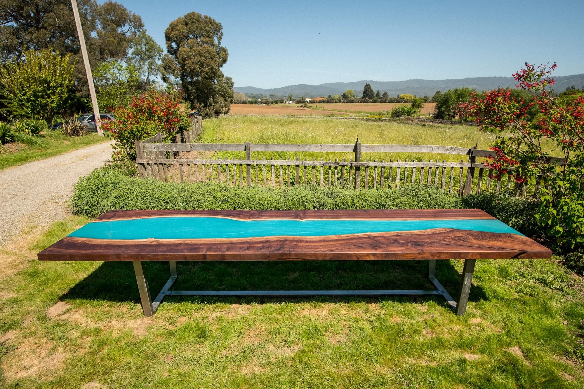 Outdoor wooden table with blue resin river running through it, placed on grass in a garden with greenery and a wooden fence, countryside landscape in the background with trees and mountains.