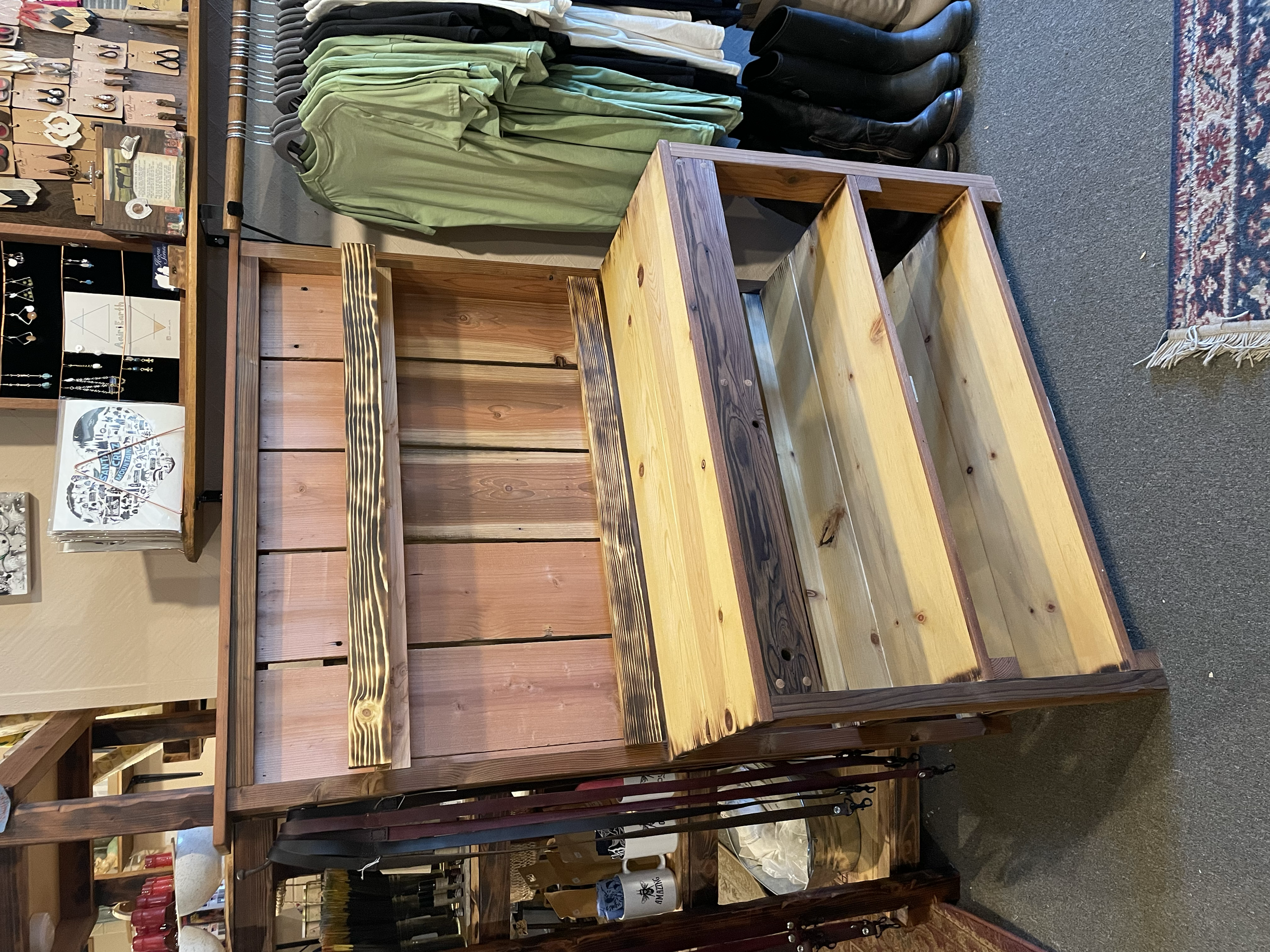 Unfinished wooden retail display fixture in a store, with shelf supports and a slatted back panel, surrounded by clothing and accessories.
