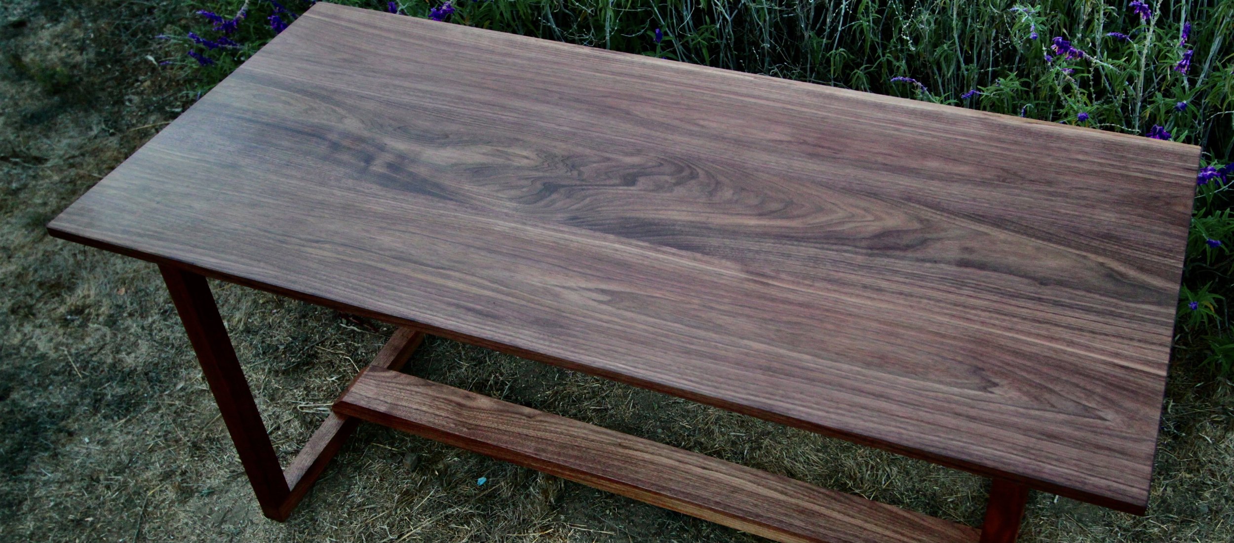 A wooden table with a slatted lower shelf, set outdoors on grass near purple flowers.