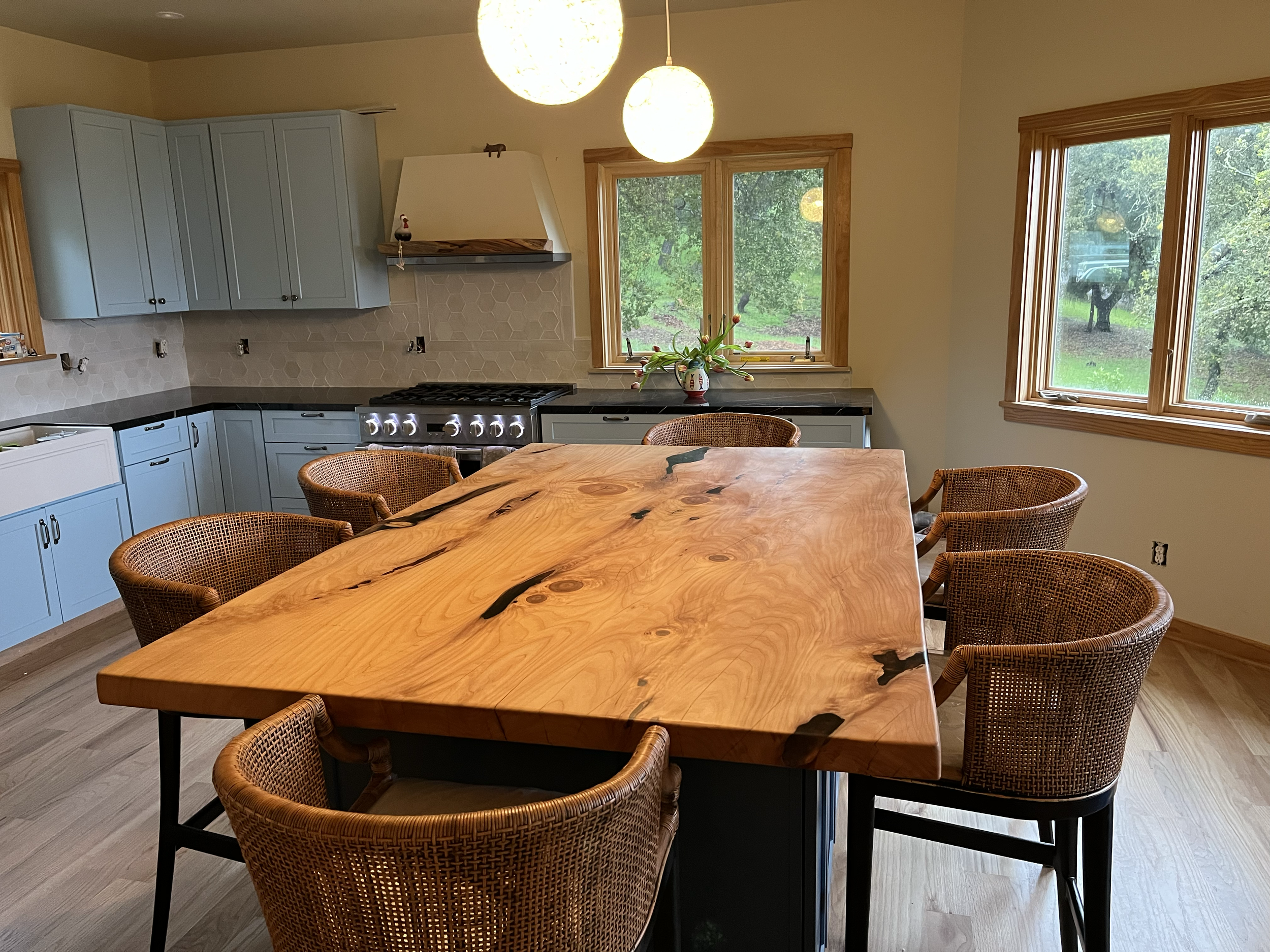 A dining area with a large, natural wood table surrounded by six wicker chairs, located in a kitchen with light blue cabinets, black countertops, and large windows overlooking a green outdoor landscape. There are hanging spherical light fixtures abov