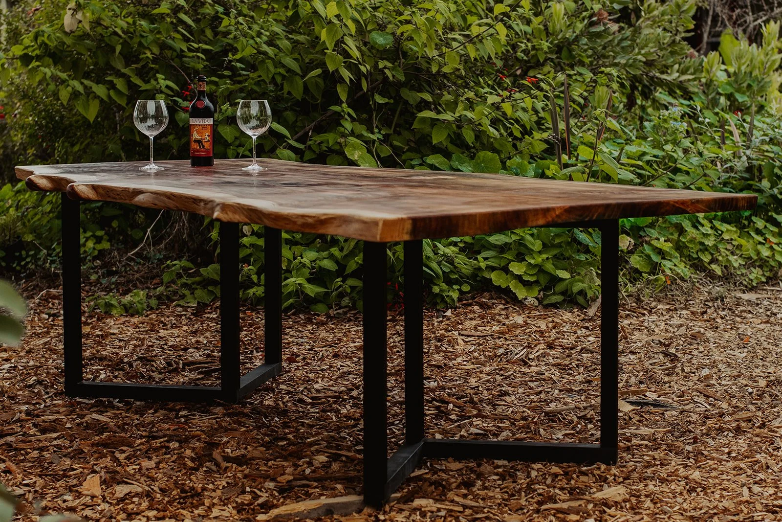 A rustic wooden outdoor table with black metal legs set on a bed of wood chips, with a bottle of red wine and two wine glasses on top, against a background of green foliage.