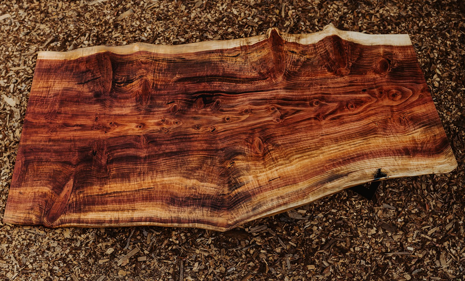 A polished, irregularly shaped wooden table with a natural edge, placed on wood chip ground.