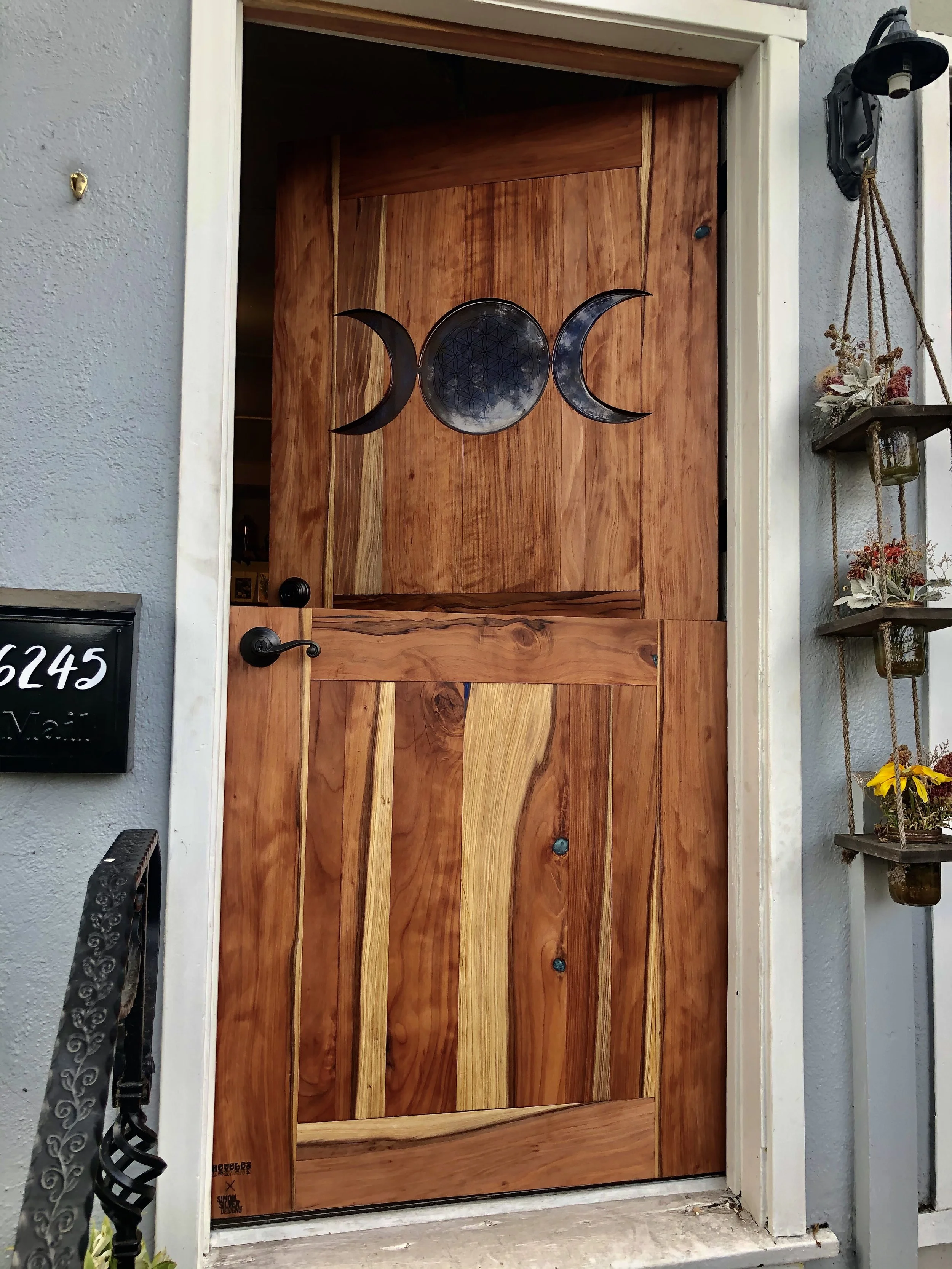 Wooden front door with a moon phases and geometric design decor on top panel, black door handle, exterior wall with hanging plant shelf, and house number 6245 on a black plaque.