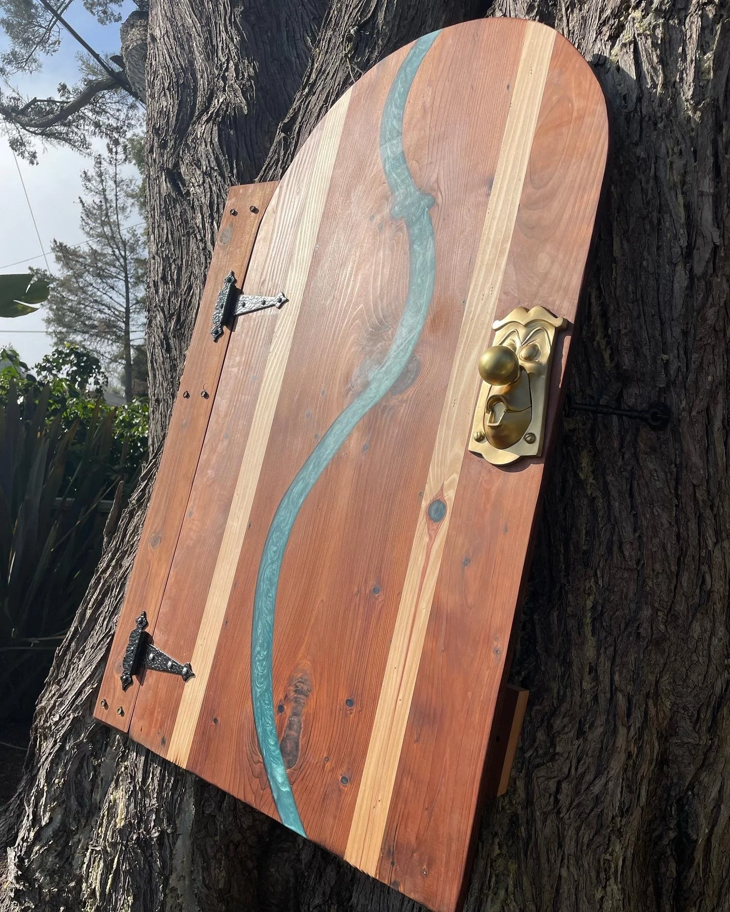 A wooden door with a painted winding river, mounted on a tree. The door has black hinges and a brass handle with a spherical knob, and is secured with a latch.
