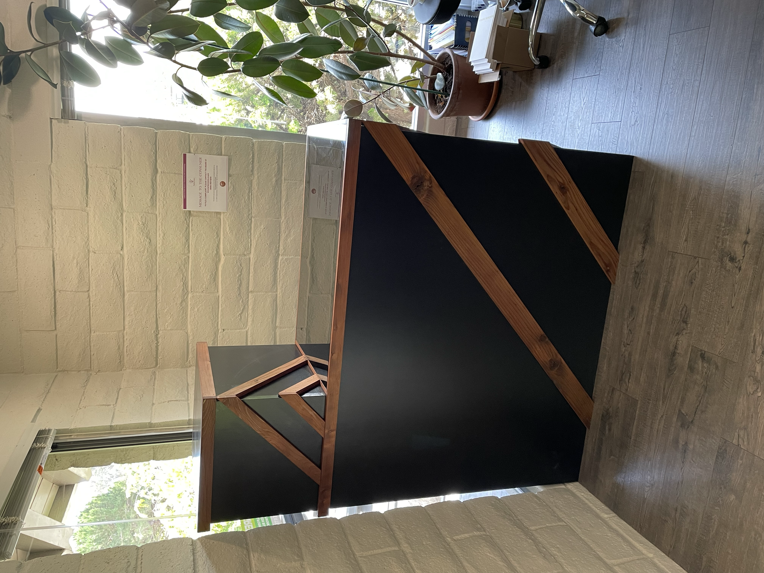 A black display stand with a wooden frame and angled design, situated near a window with potted plants and informational signs nearby.